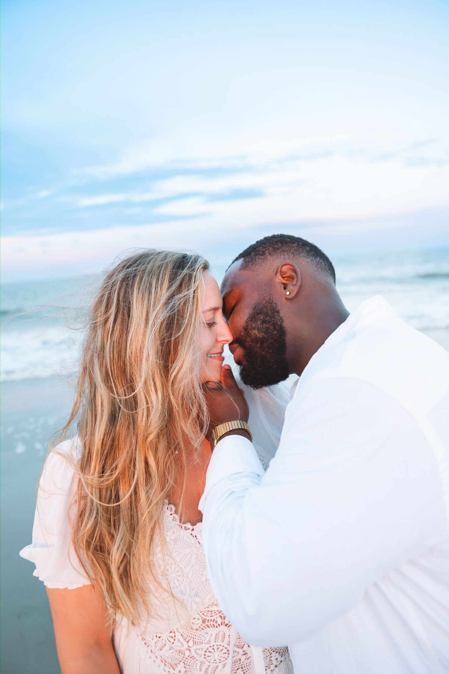 Interracial couple sharing a tender moment on a beach at dusk with soft blue sky behind them.