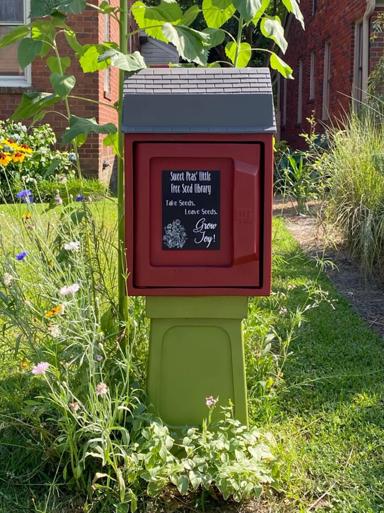Green Little Free Seed Library box mounted on post surrounded by wildflowers outside a brick building.