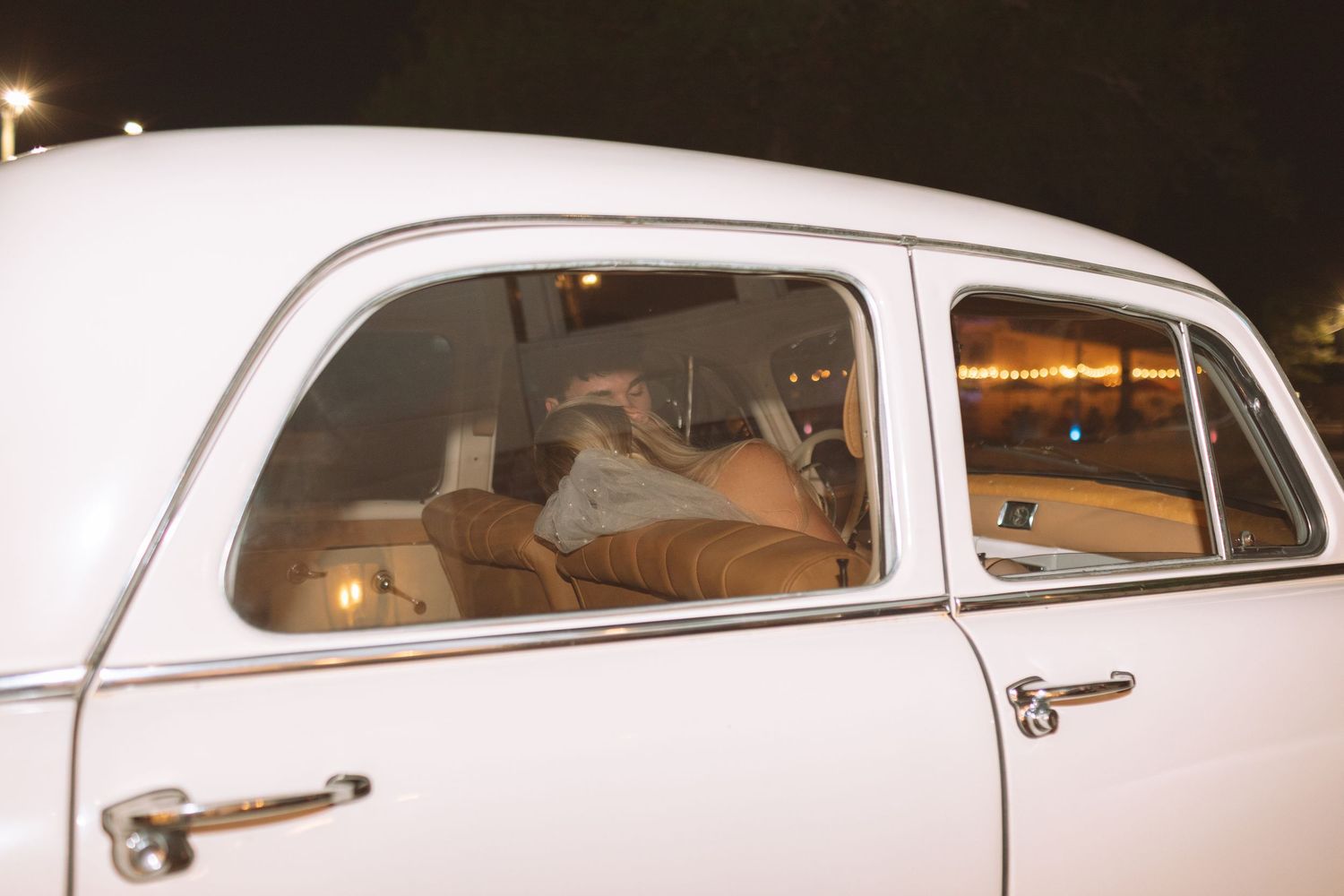 Newlyweds sharing a kiss inside a classic vintage white getaway car at night.