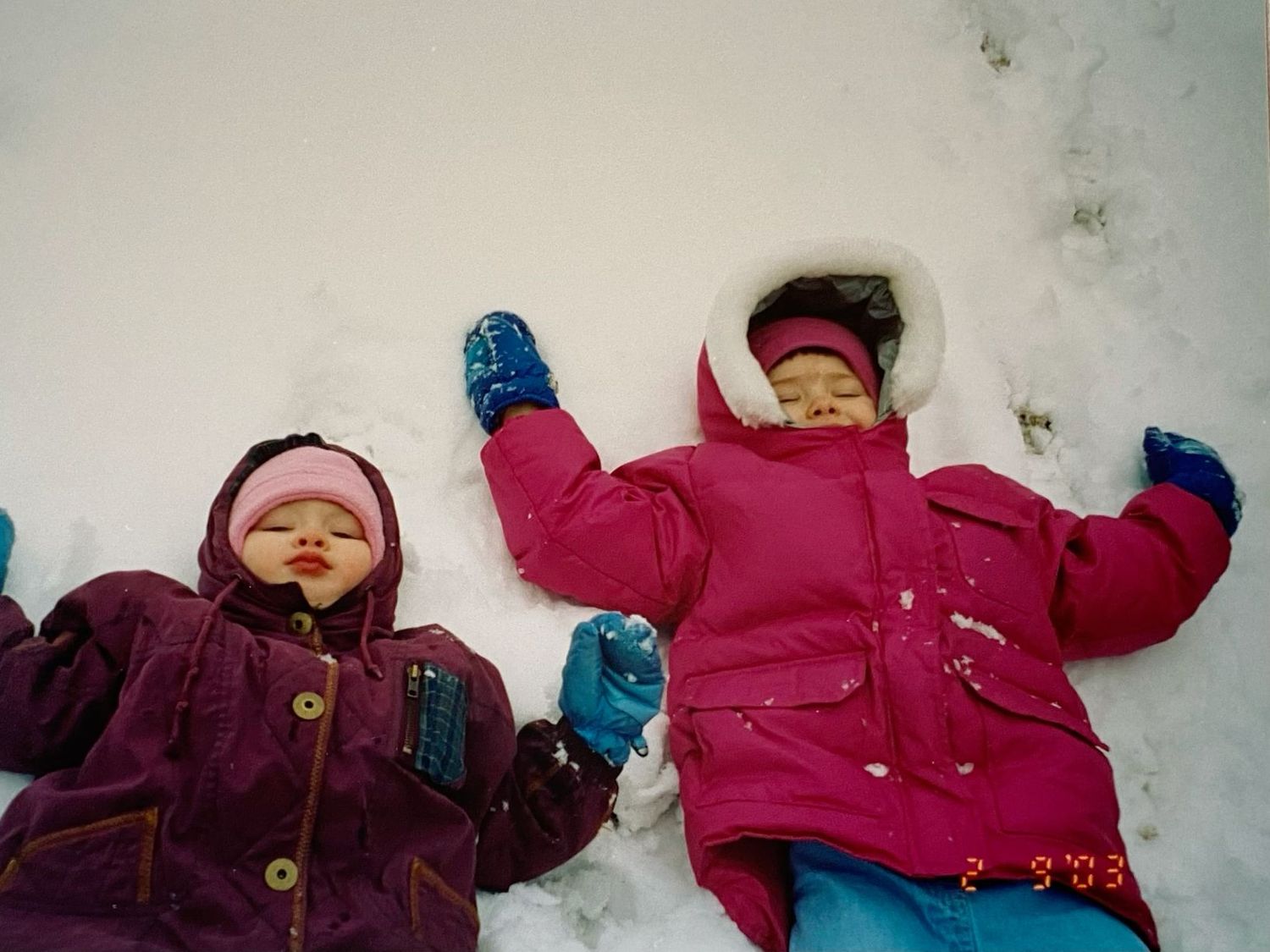 Two young toddlers in colorful winter jackets making snow angels in fresh white snow outdoors.