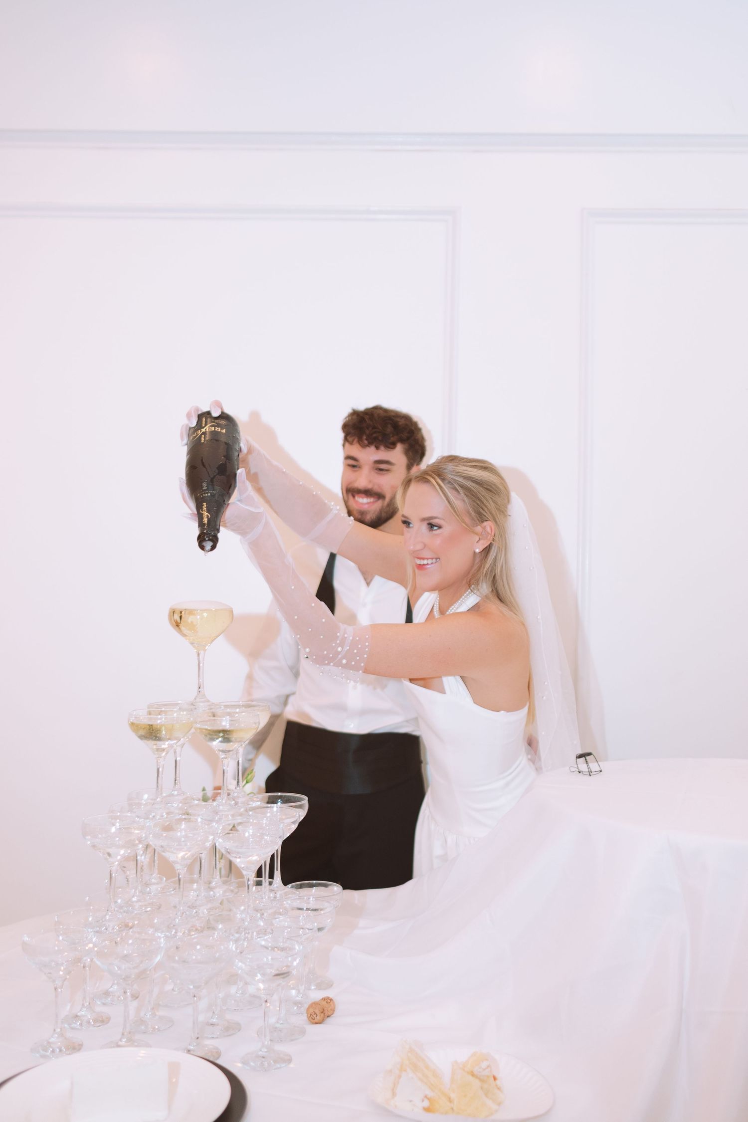 Newlyweds pouring champagne into elegant glass tower at wedding reception.