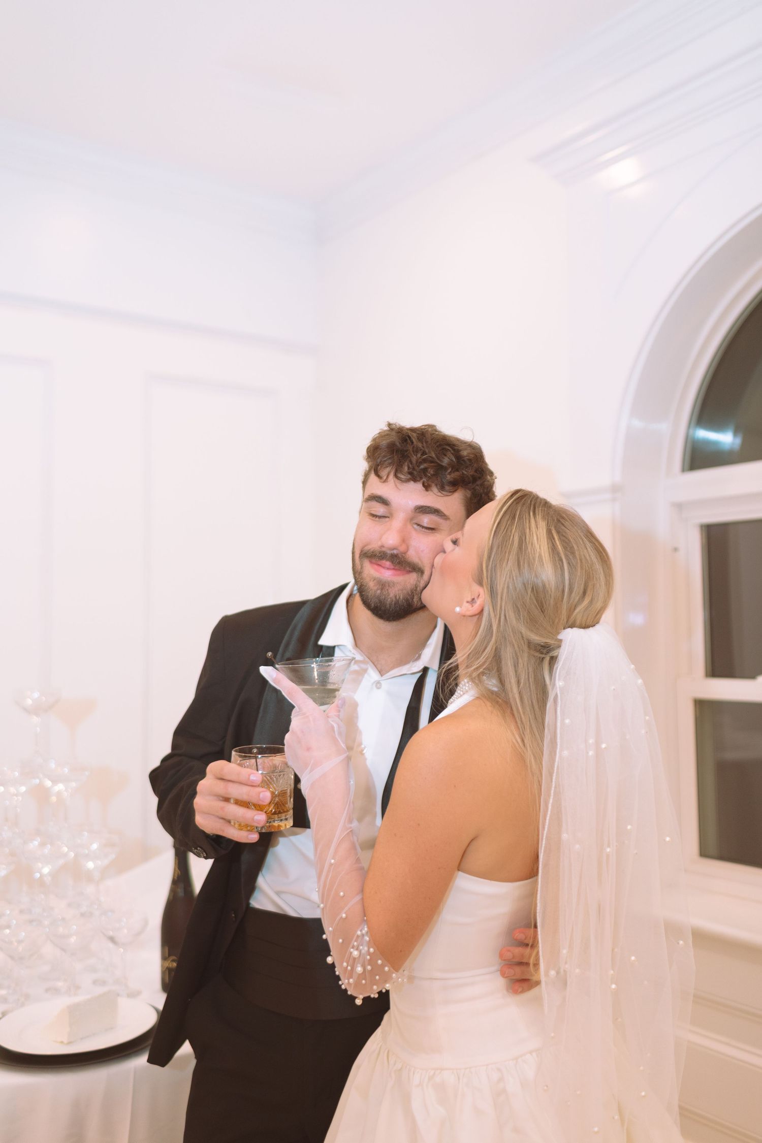 Bride kissing groom on the cheek while he holds cocktails at their wedding reception.