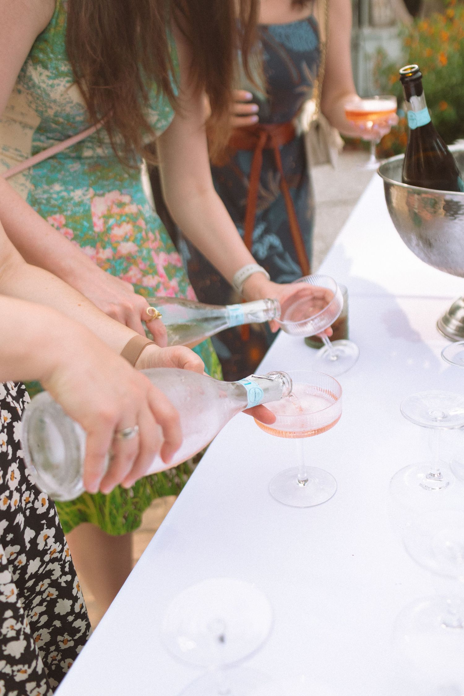 Wedding guests pour pink champagne into elegant coupe glasses at outdoor reception drink station.
