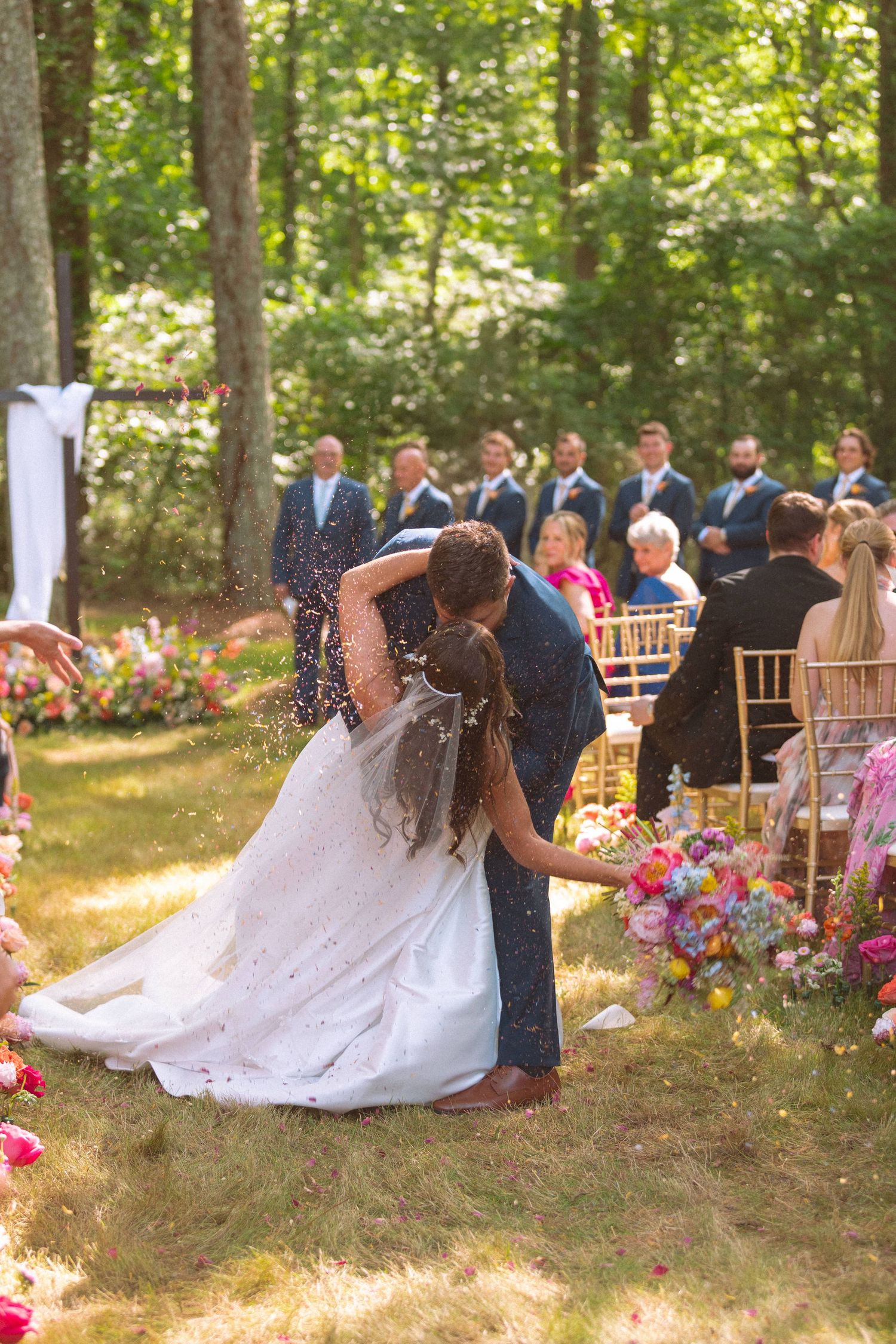 Groom dips bride for first kiss during colorful outdoor garden wedding ceremony with guests.