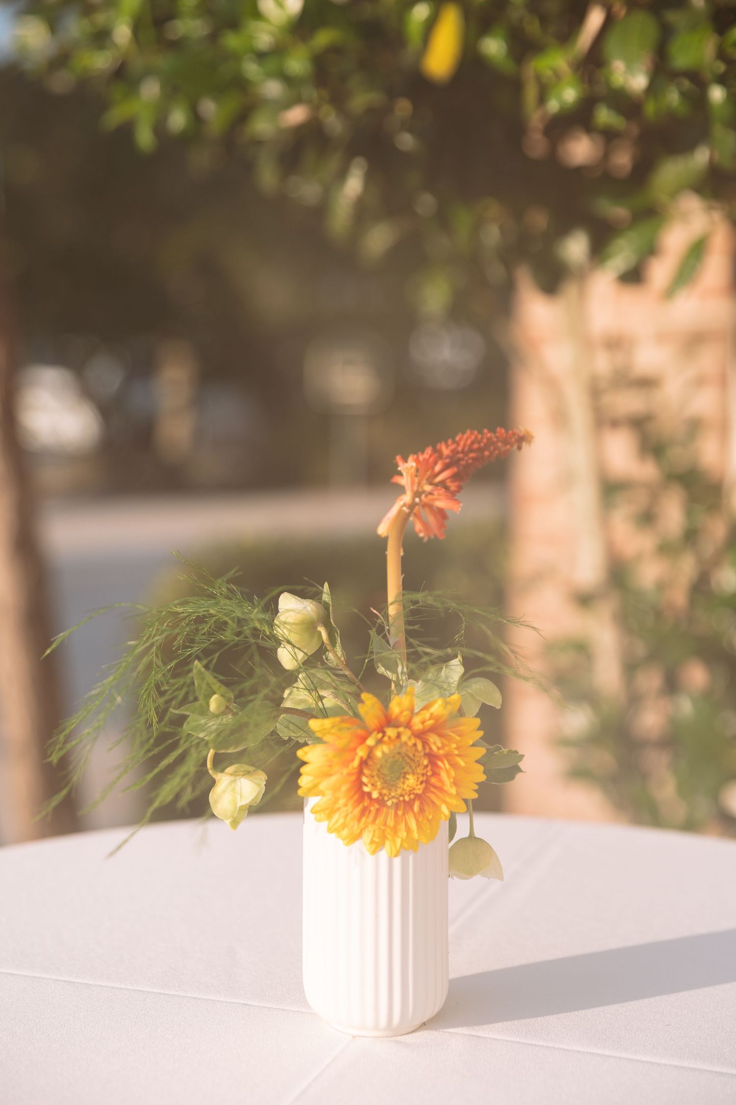 Sunflower and orange bloom arrangement in white ribbed vase centerpiece on white table at outdoor wedding.