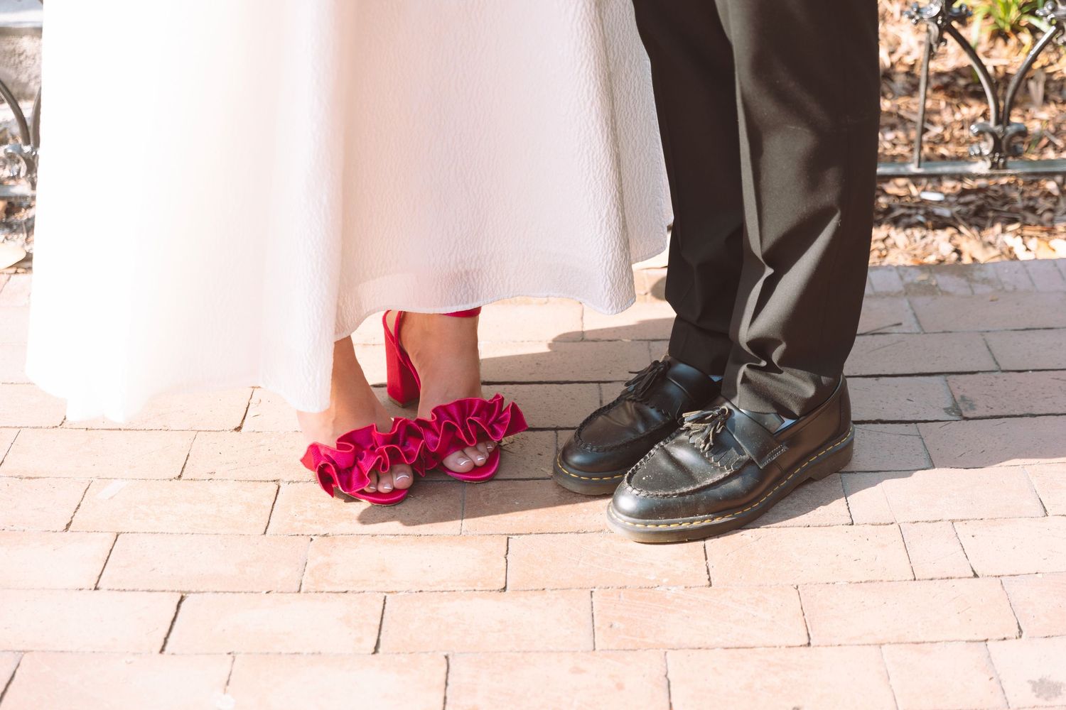 Close-up of bride in pink heels and groom in black platform shoes standing on brick pavement.
