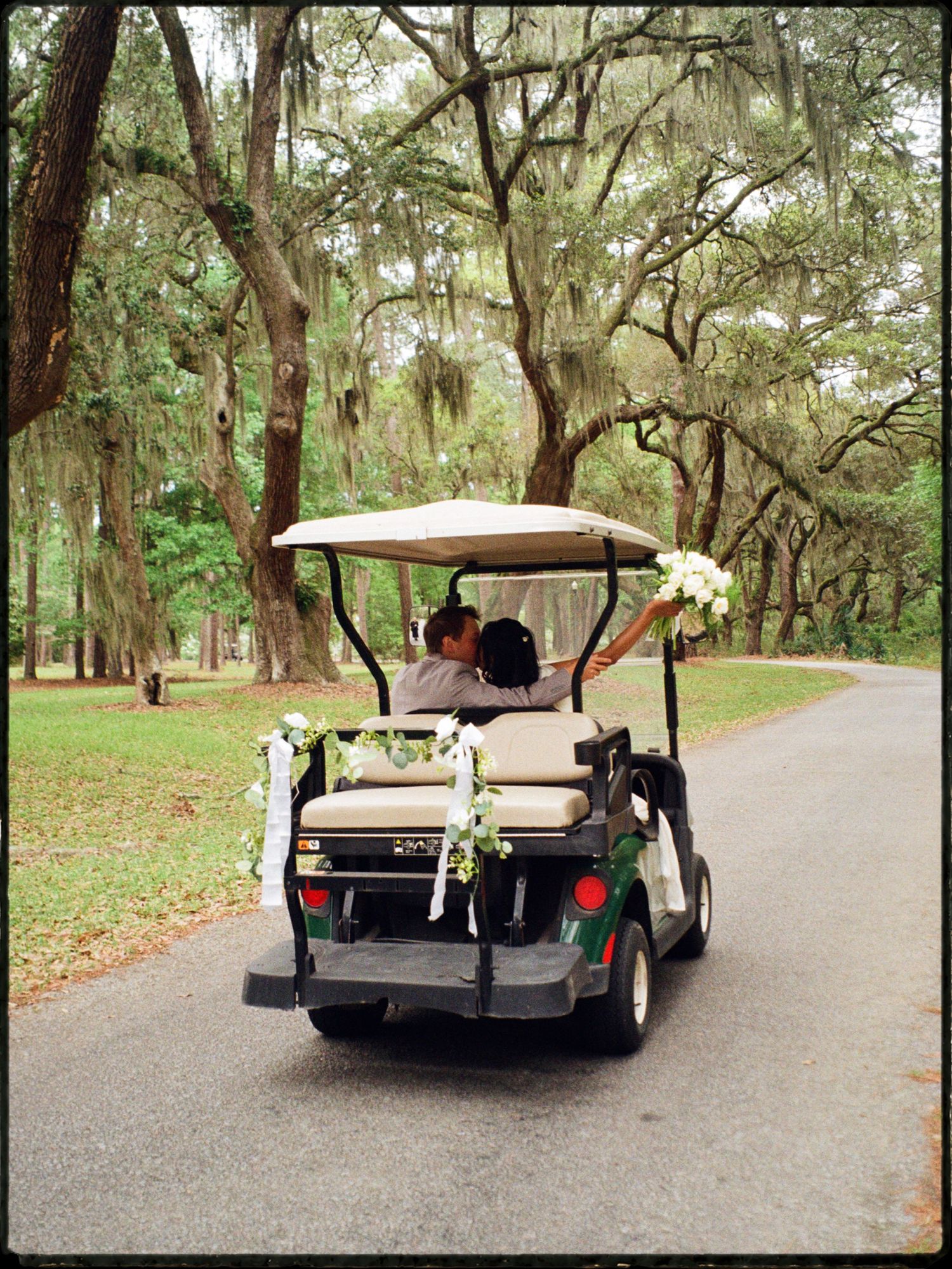 Newlyweds riding a flower-decorated golf cart down a tree-lined path draped with Spanish moss.