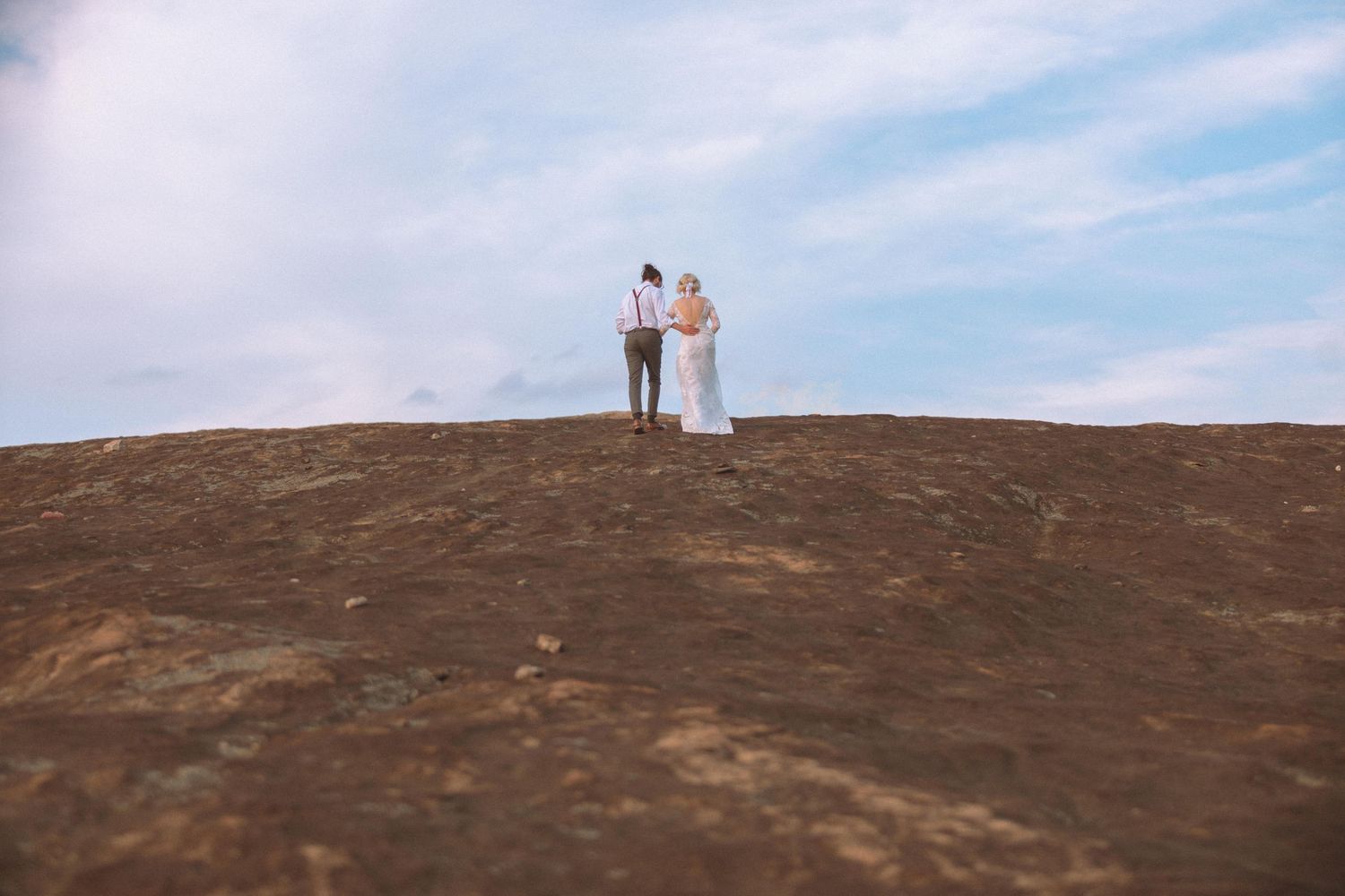 Couple in wedding attire standing atop a rocky hill under a blue sky.