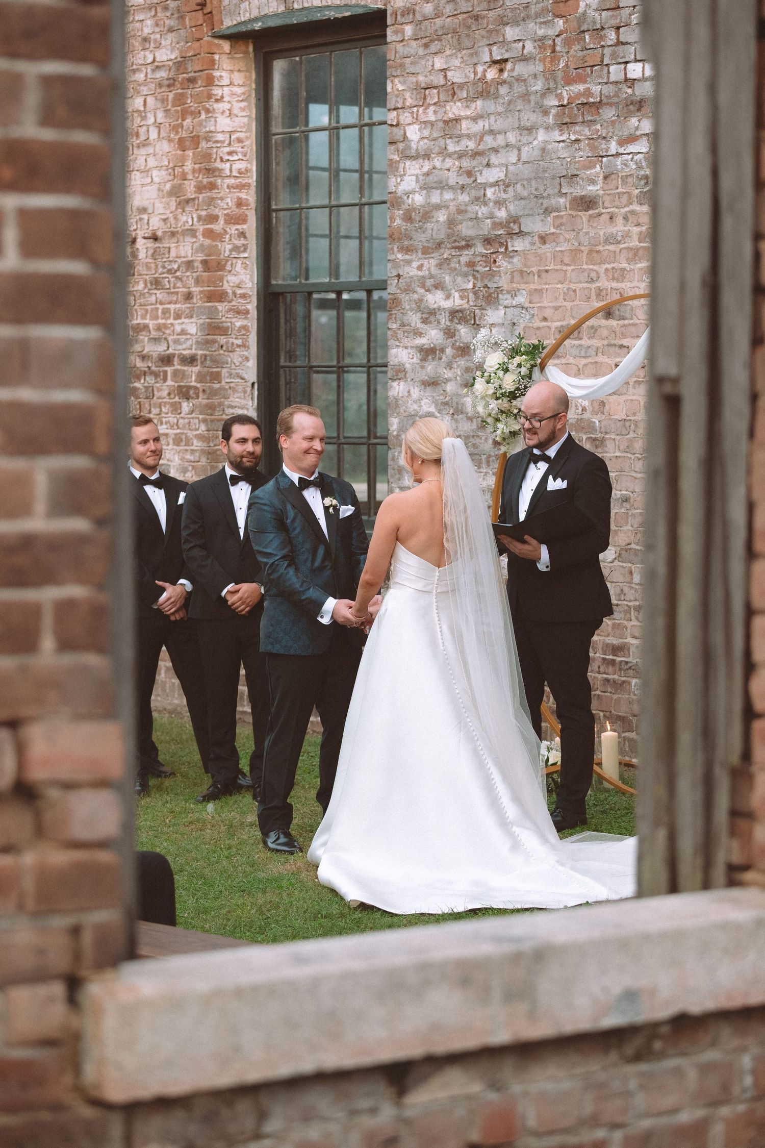 Bride and groom exchange vows outdoors against a rustic brick wall with groomsmen standing nearby.