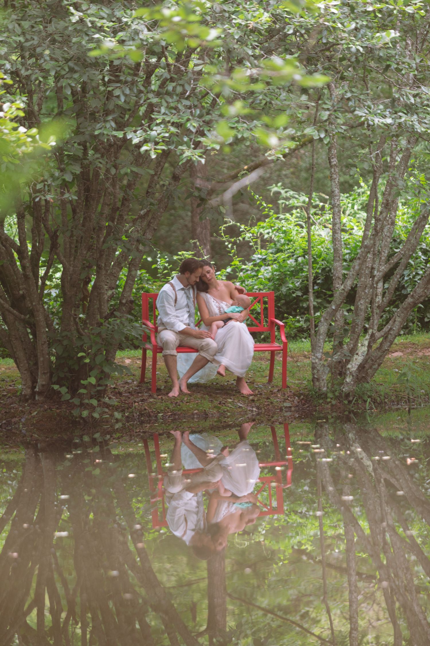 Wedding couple sits on red bench kissing, reflected in still pond amid lush trees.