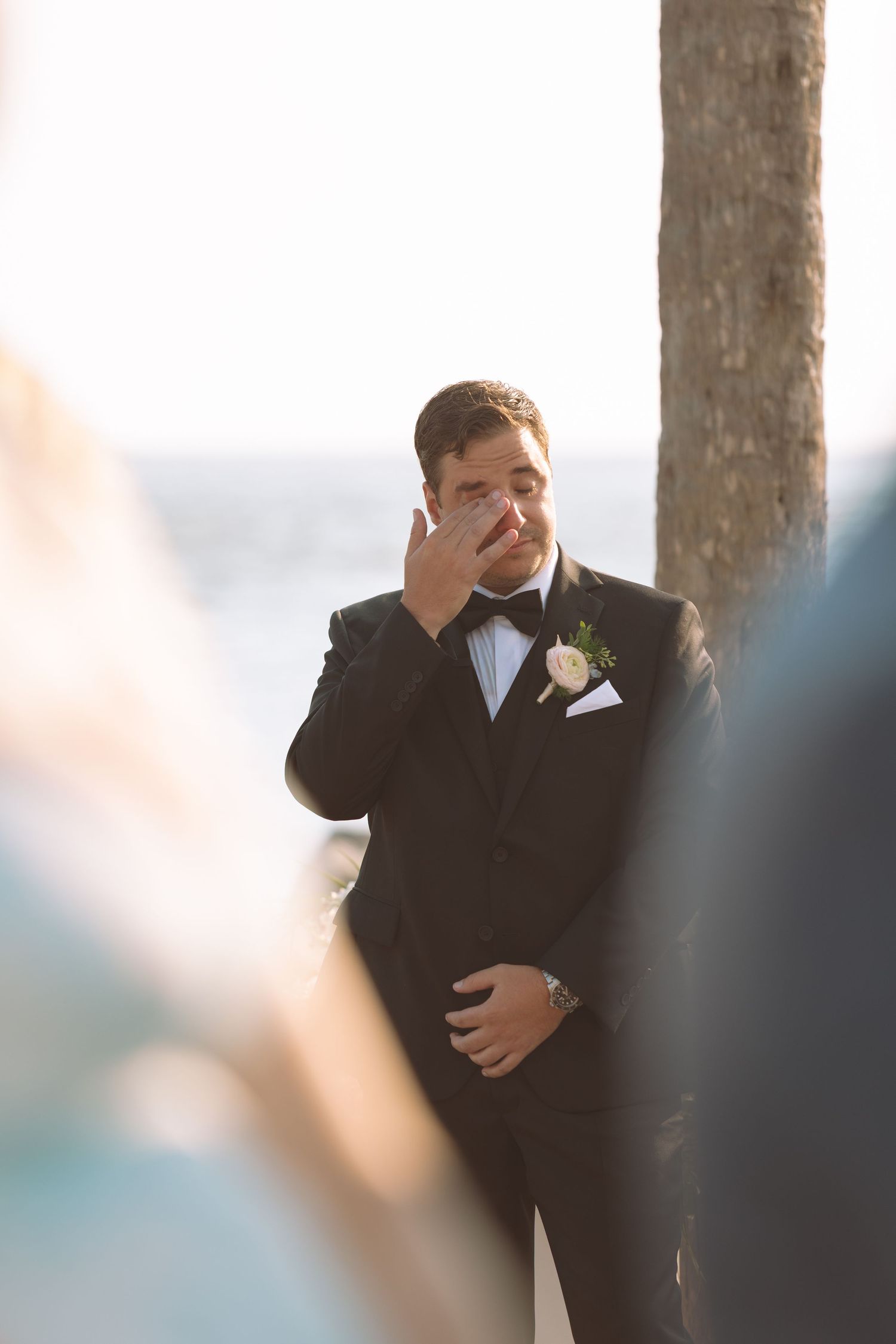 Emotional groom in black tuxedo wipes tears while waiting at the altar during an outdoor wedding ceremony.