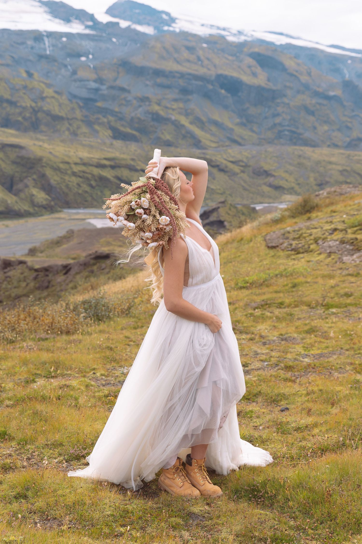 Bride in white tulle gown and boots holding floral bouquet on scenic mountain hillside in Iceland.