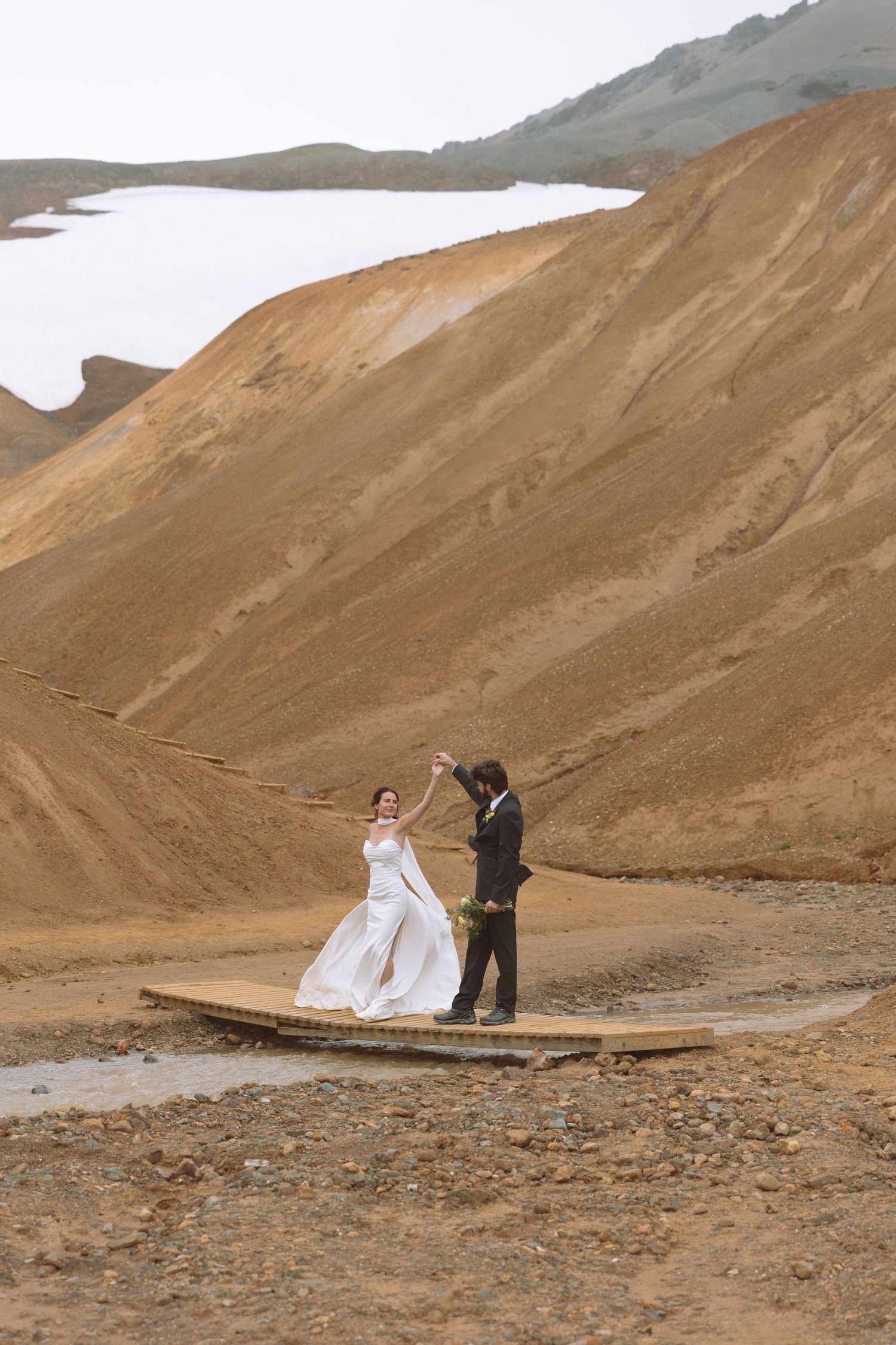 Wedding couple dances on a wooden bridge surrounded by Iceland's stunning golden-brown volcanic landscape.