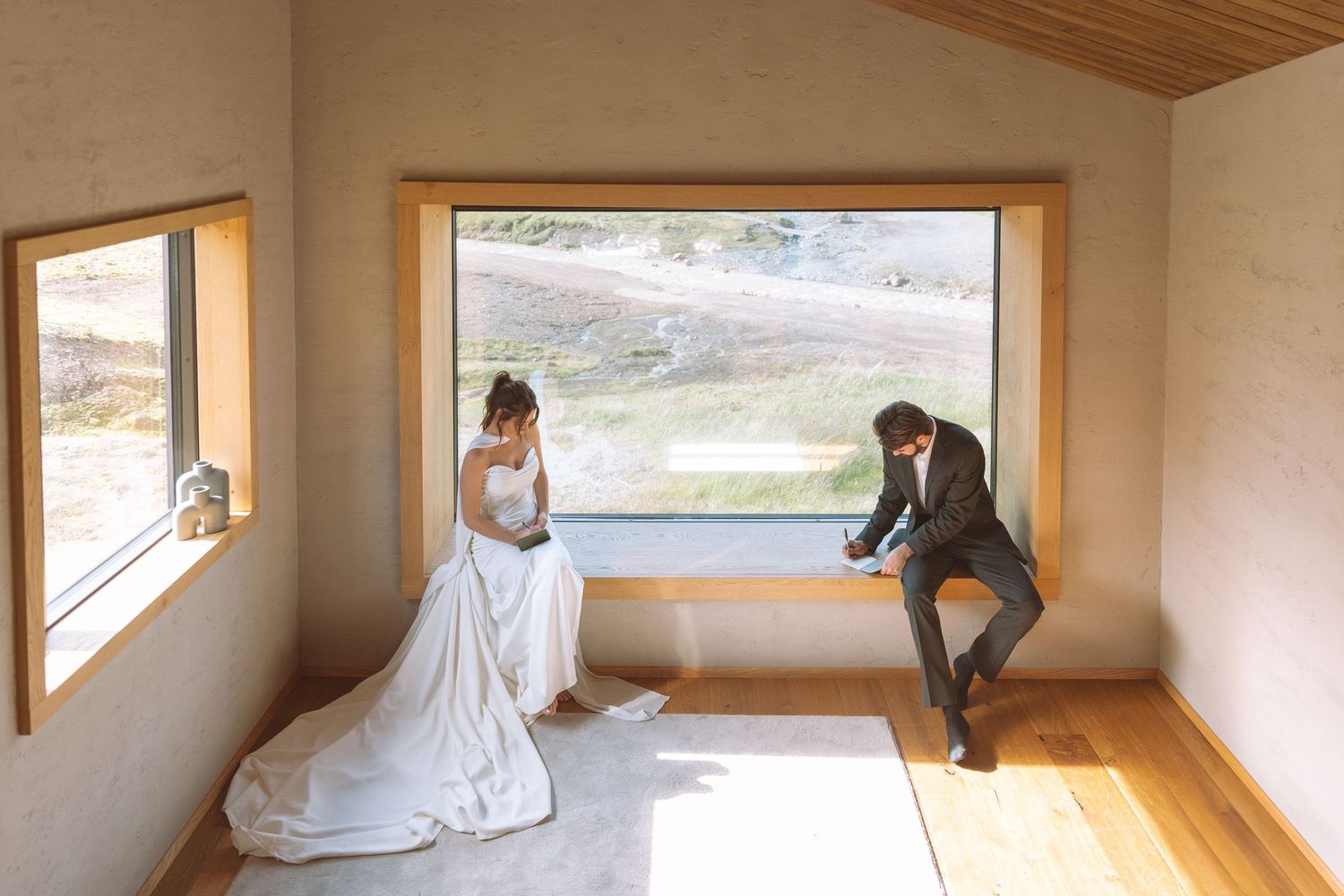 A bride and groom sit separately by a large panoramic window in a modern wooden interior space.