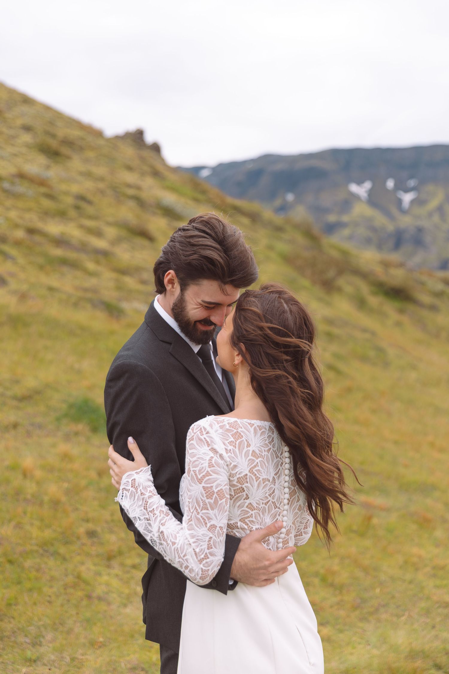 Groom leans in to kiss bride while embracing on a lush green mountain hillside.
