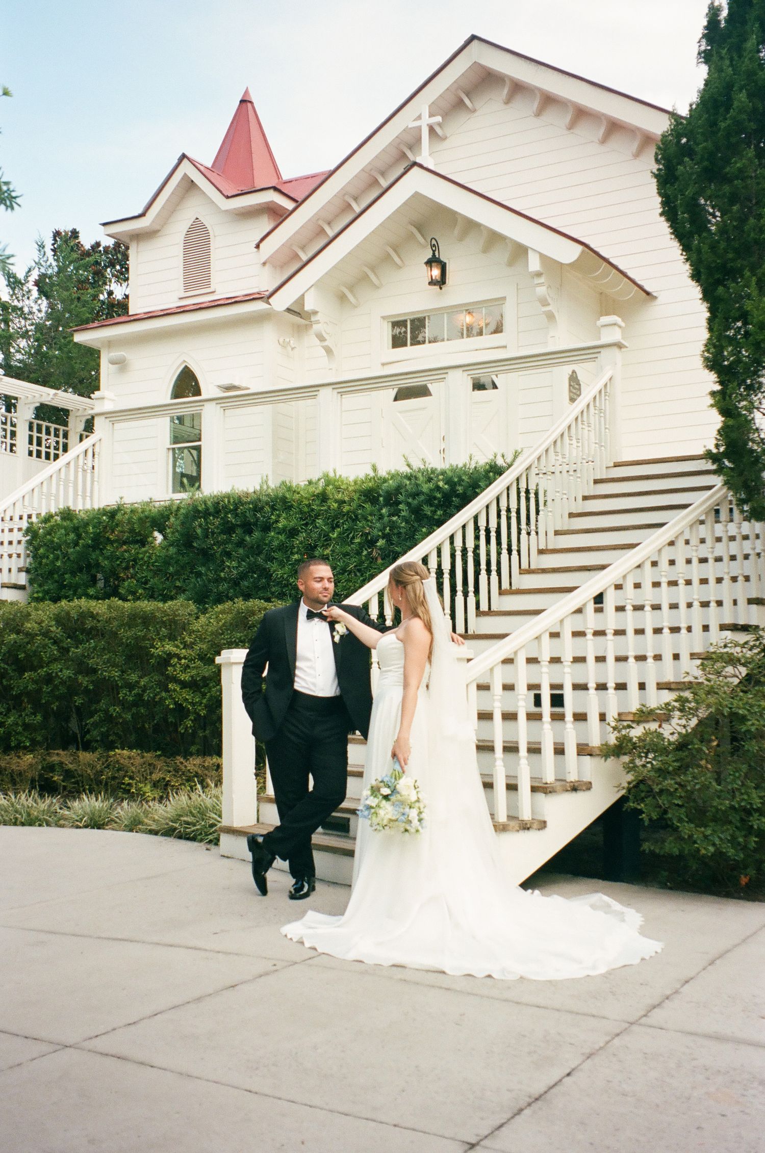 Bride and groom posing in front of a charming white wedding chapel with a red steeple and grand staircase.