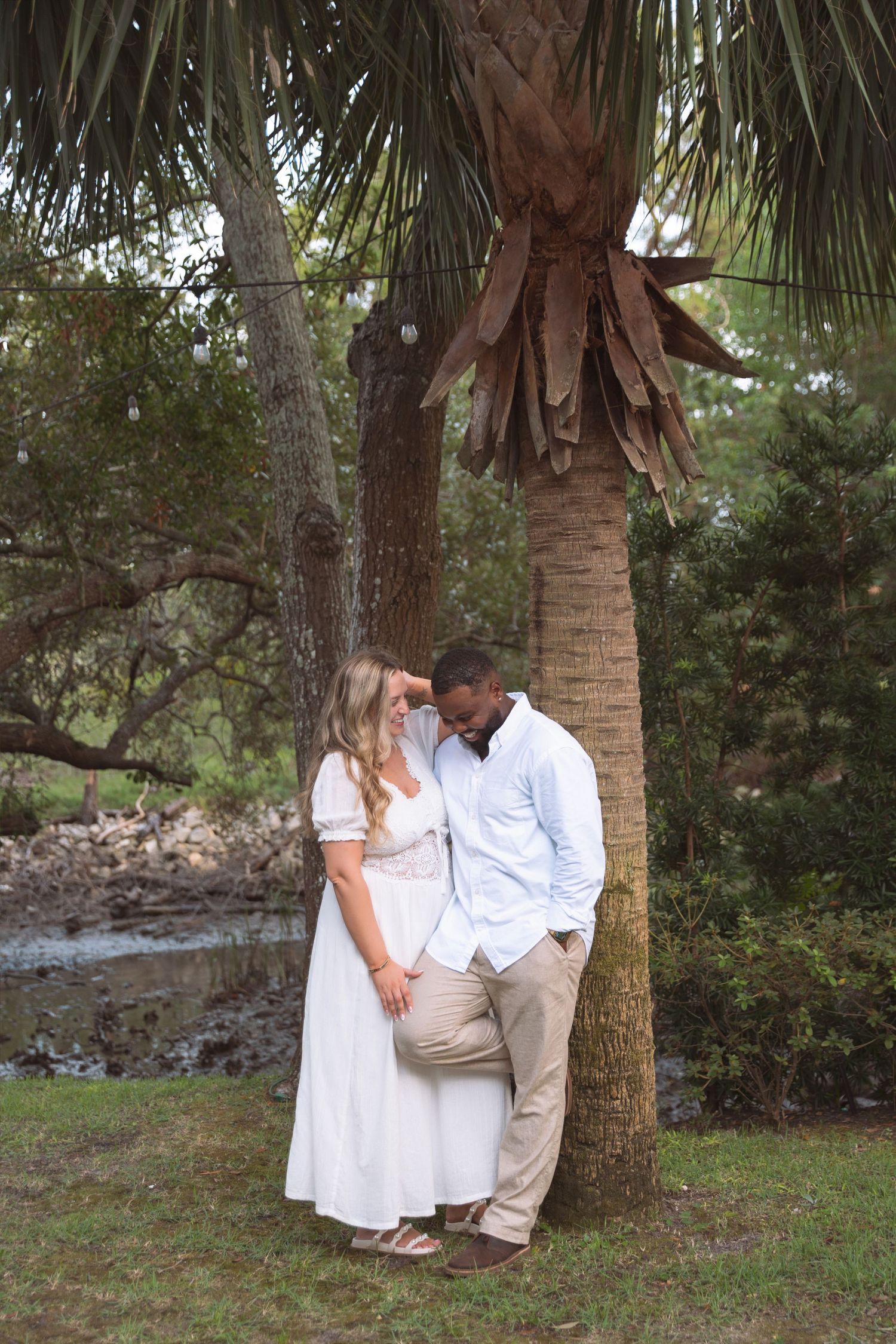 Interracial couple sharing a tender moment leaning against a palm tree in a lush green outdoor setting.