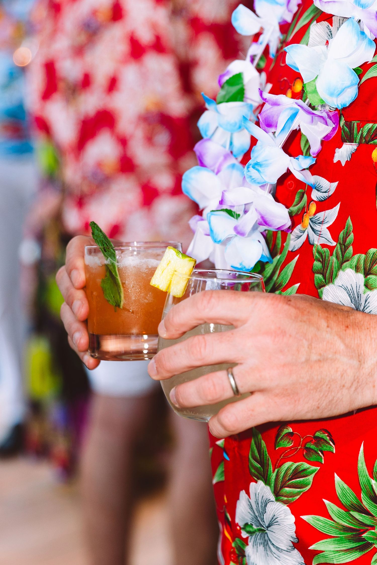 Man in red Hawaiian shirt and lei holding tropical cocktail garnished with pineapple and mint.