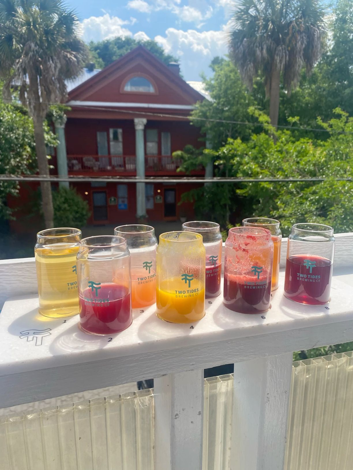 Colorful layered cocktails lined up on white railing with tropical house in background.