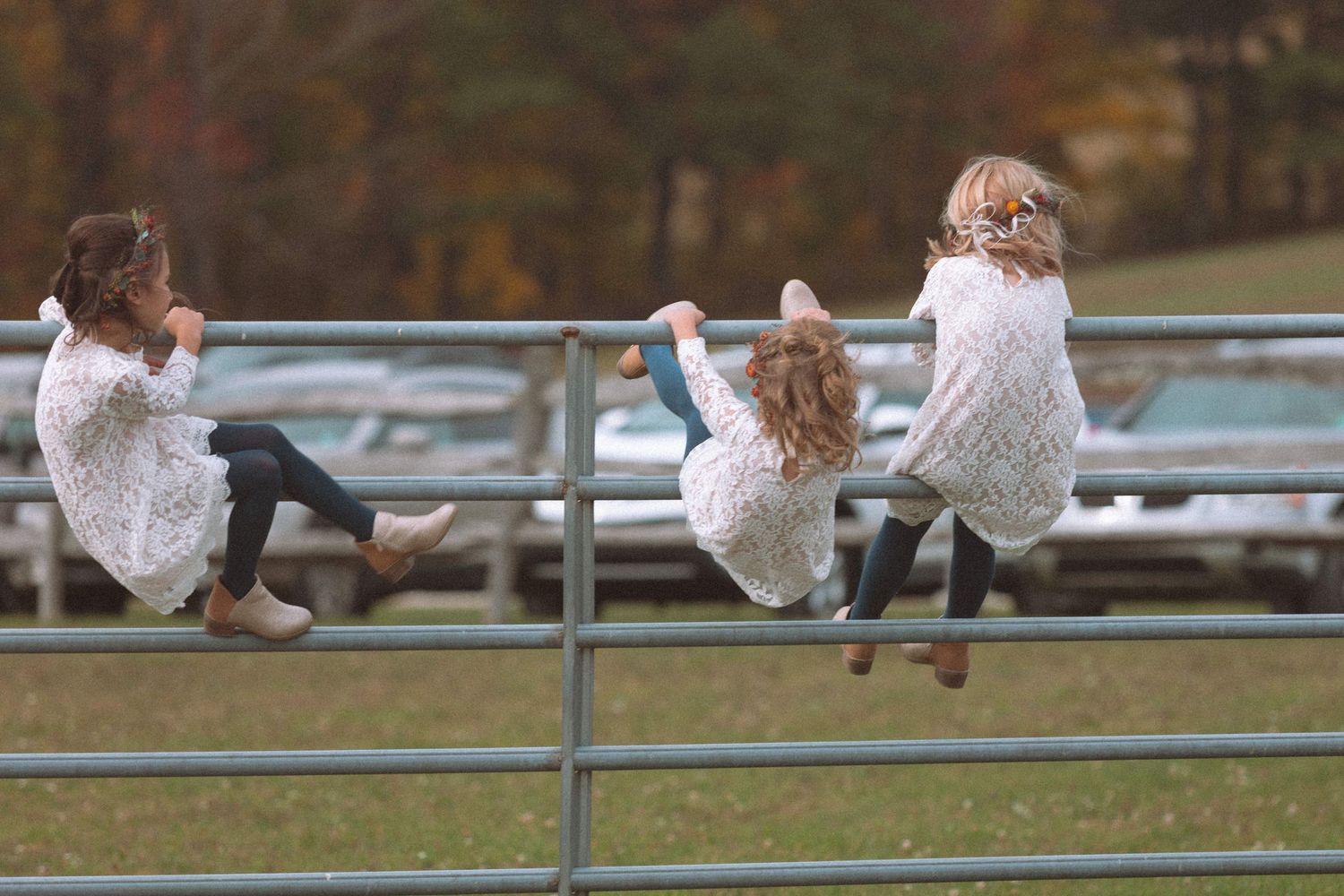 Three young girls in white dresses hanging on metal fence railing at outdoor wedding.