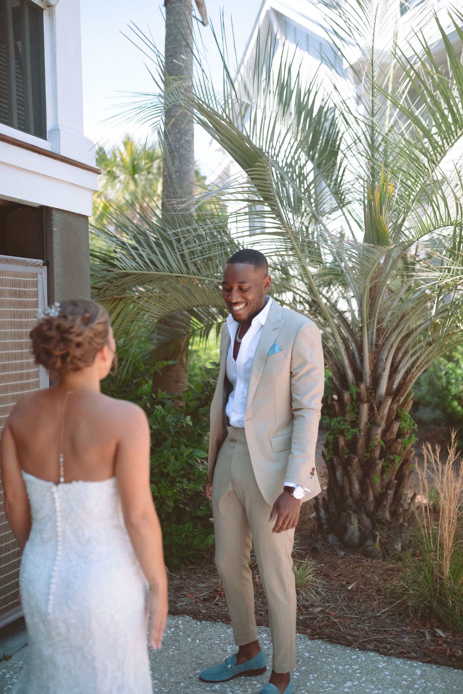 Groom in tan suit smiles seeing bride in white lace wedding gown for first look outdoors near palm trees.