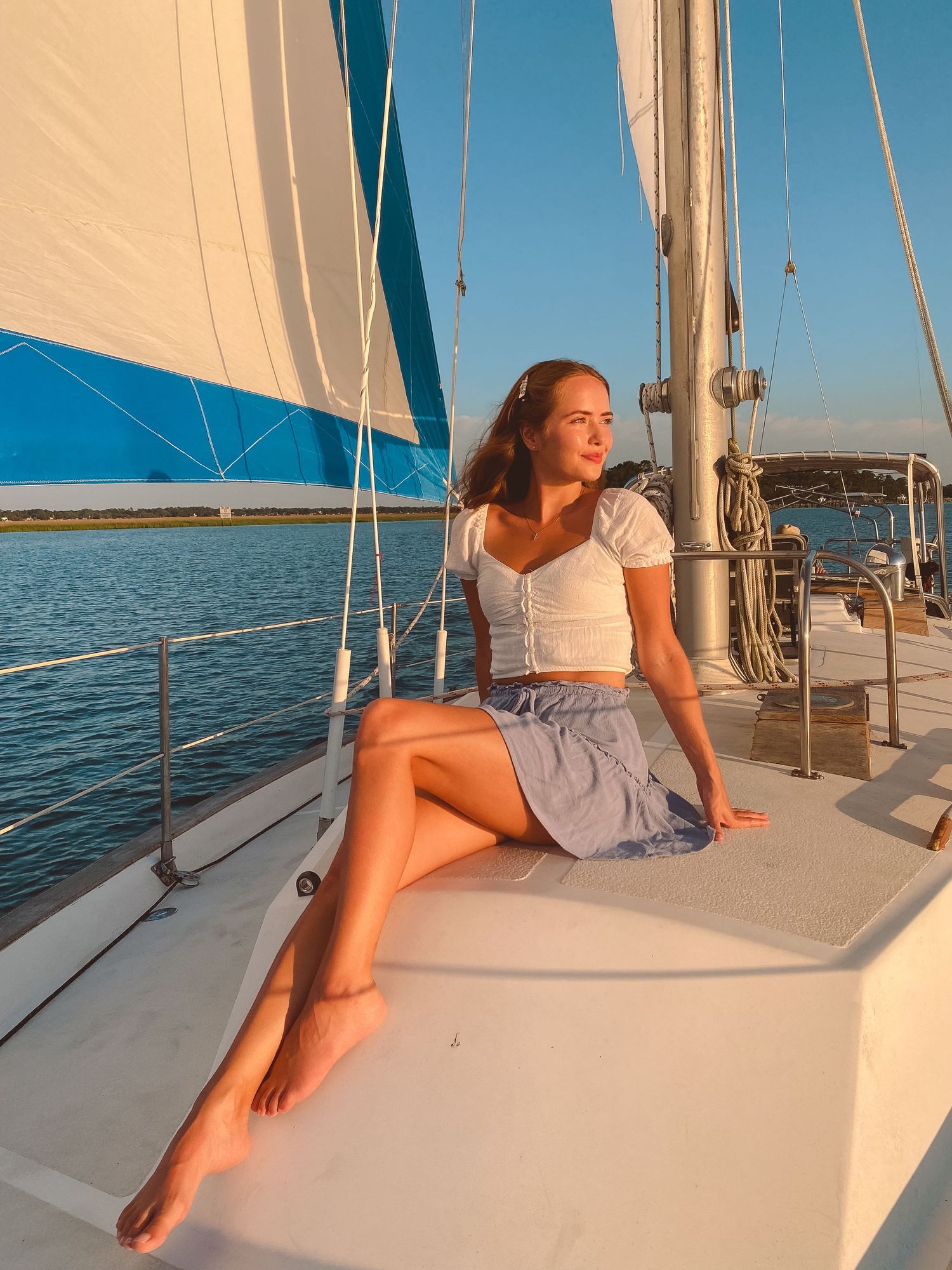 Young woman in white top and blue skirt sitting barefoot on sailboat deck at golden hour sunset.