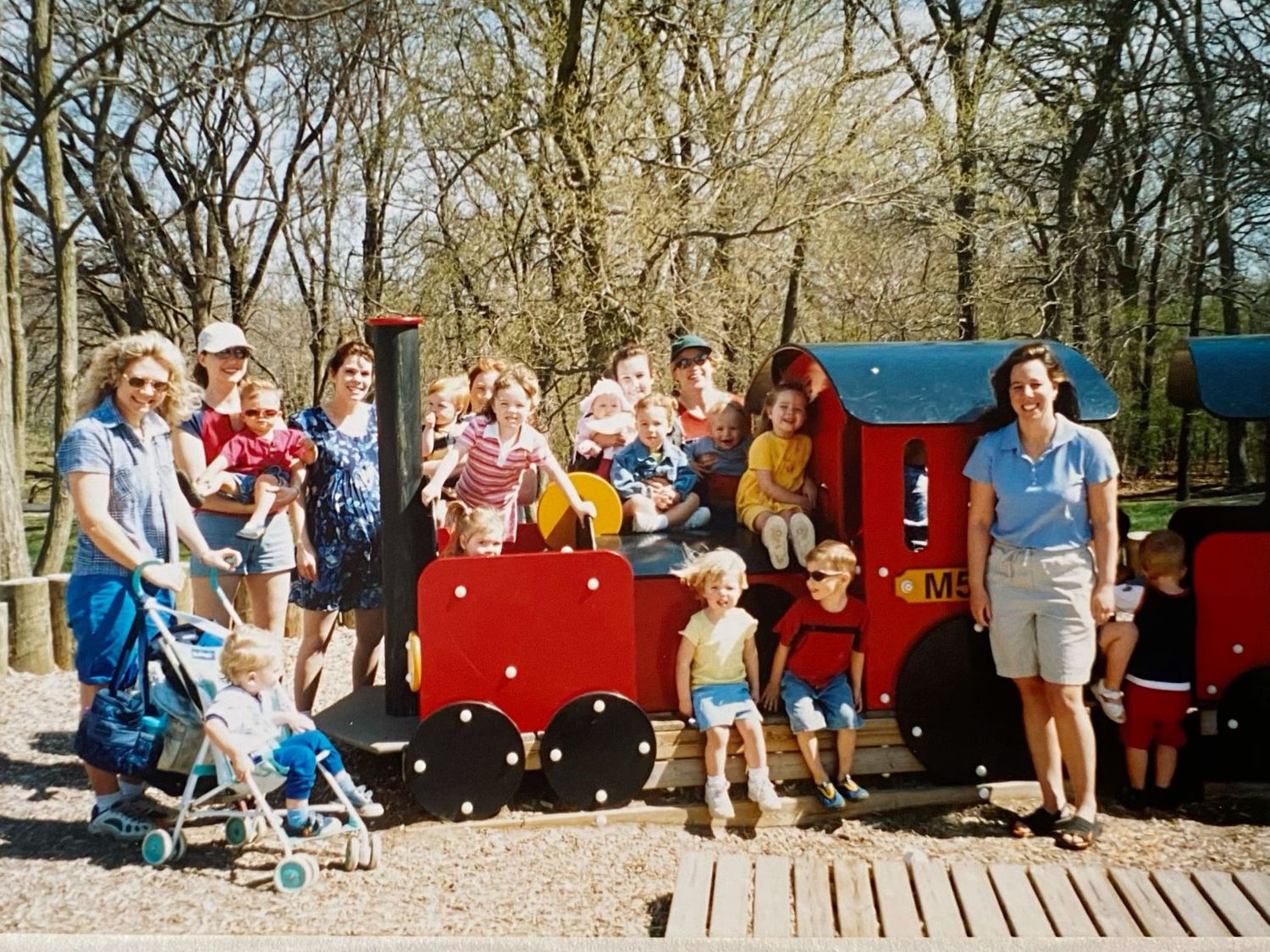 Group of families with young children posing on a colorful red and blue wooden train playground structure outdoors.