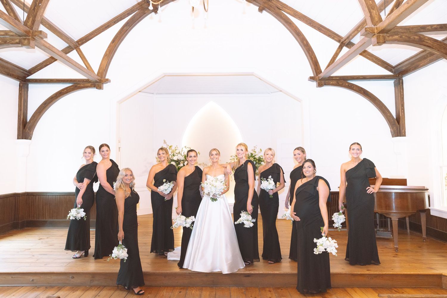 Bride in white gown poses with bridesmaids in black dresses holding white bouquets inside elegant chapel venue.