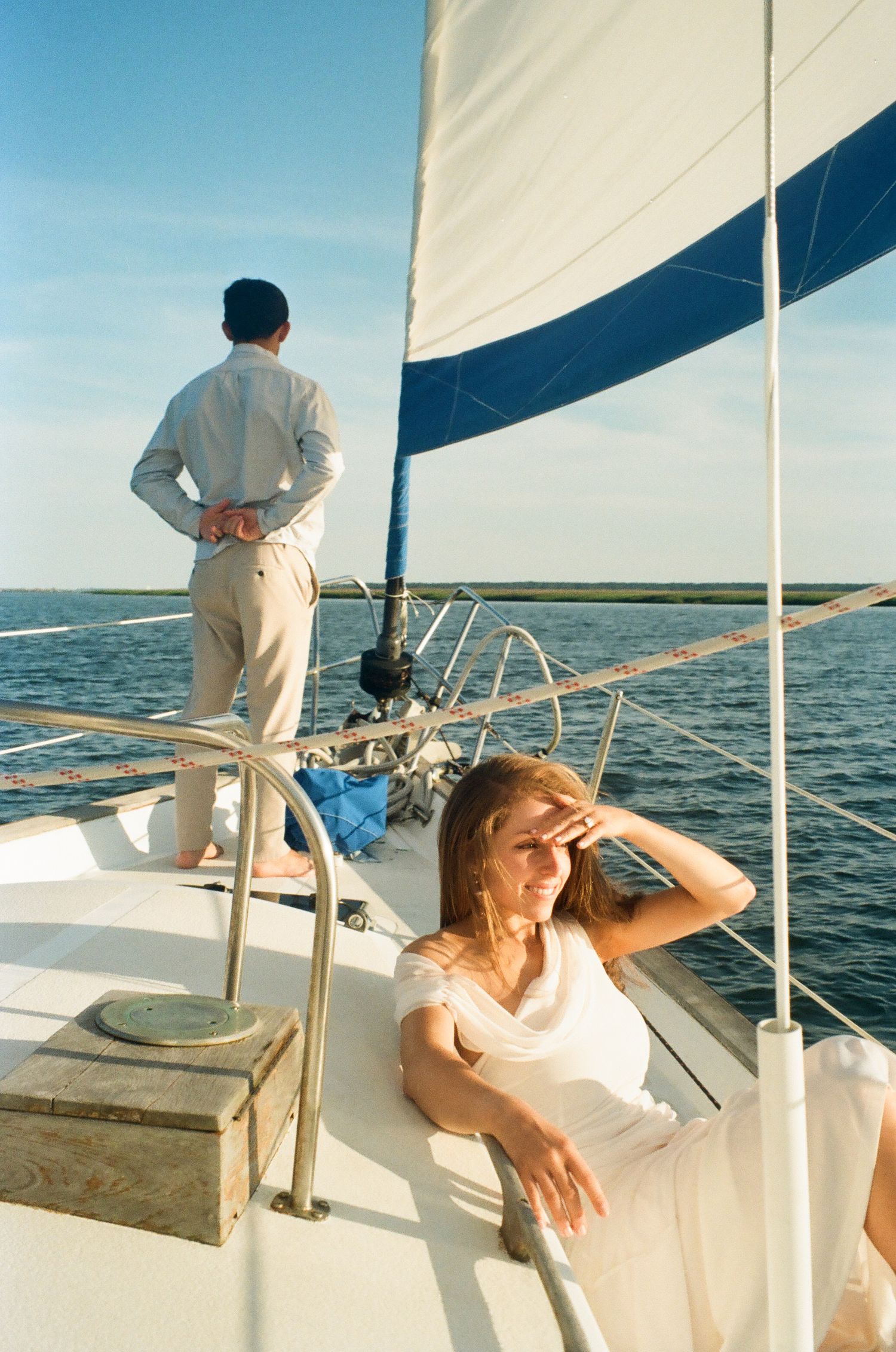 Woman relaxing on sailboat deck shielding eyes from sun while man stands at stern overlooking calm blue ocean.