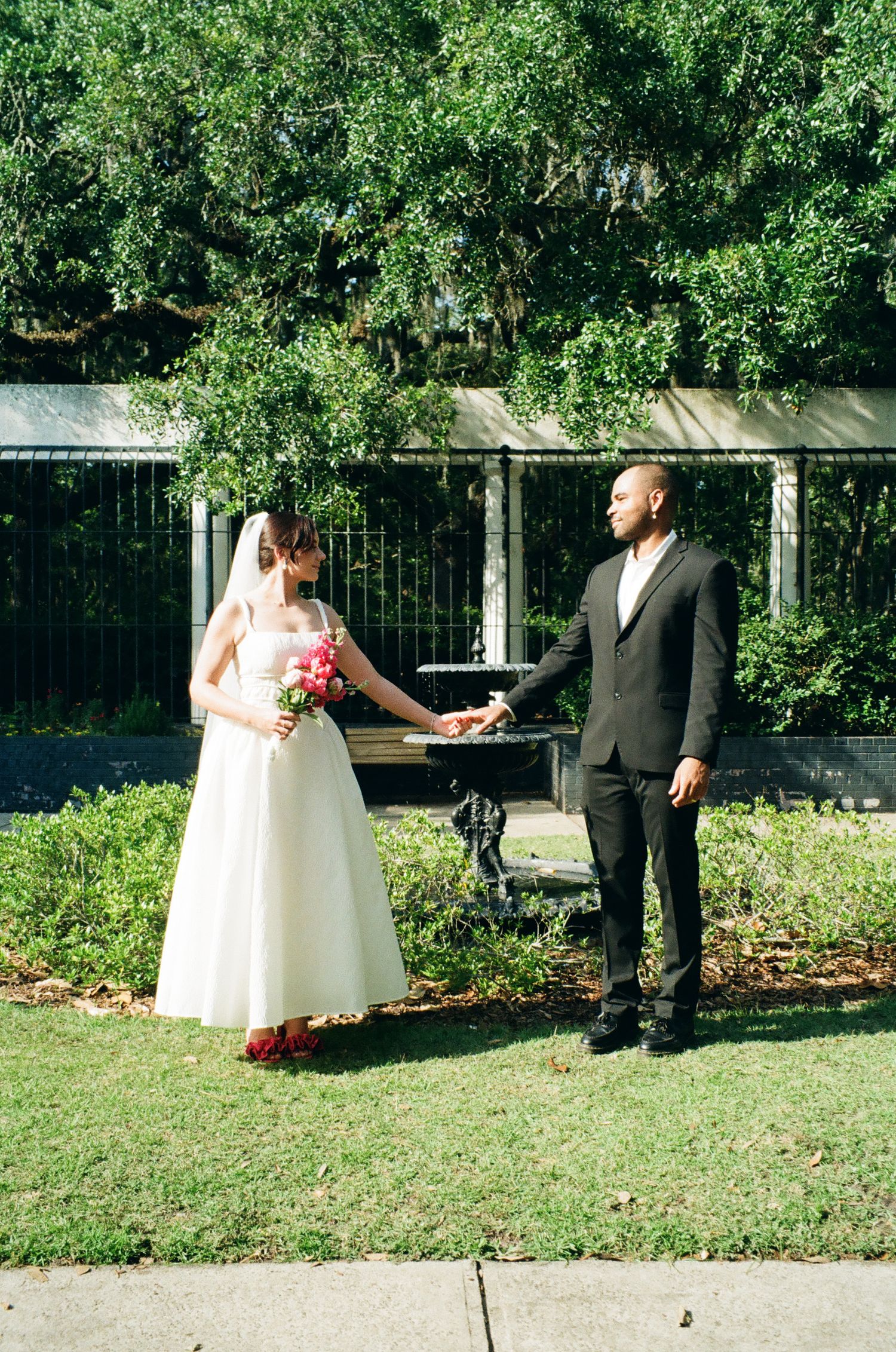 Bride in white gown holding pink bouquet and groom in dark suit hold hands outdoors in a garden setting.