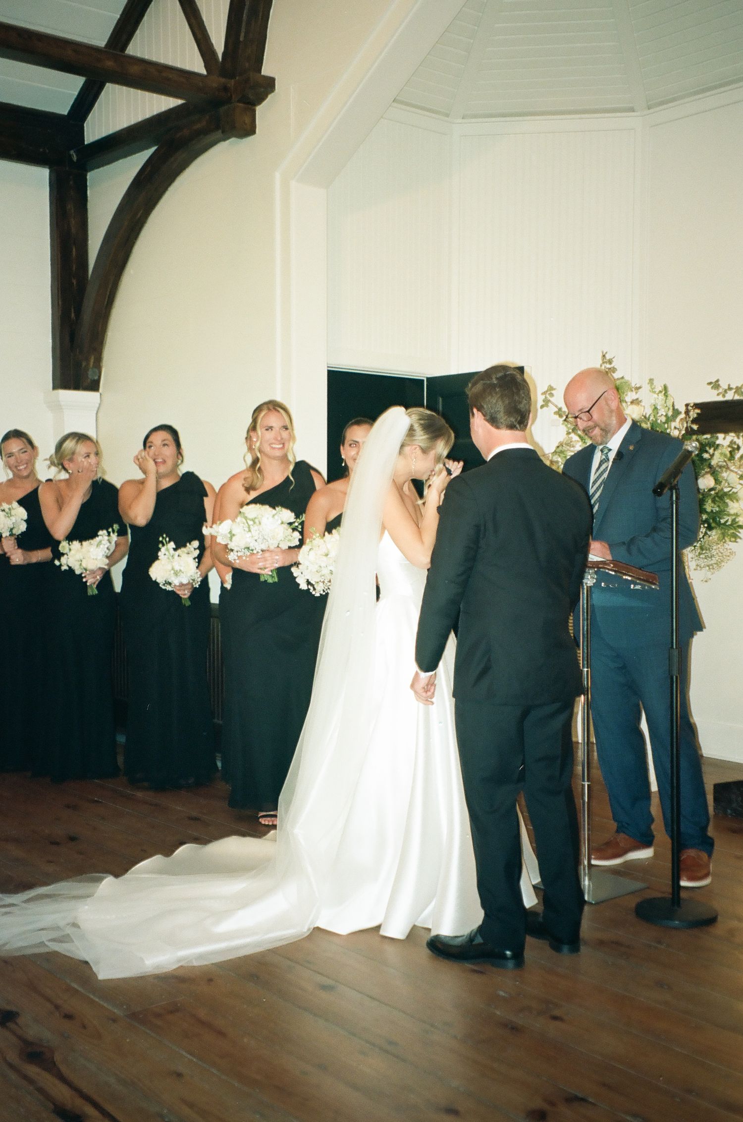 Bride and groom share a kiss at altar during indoor wedding ceremony with bridesmaids in black dresses.