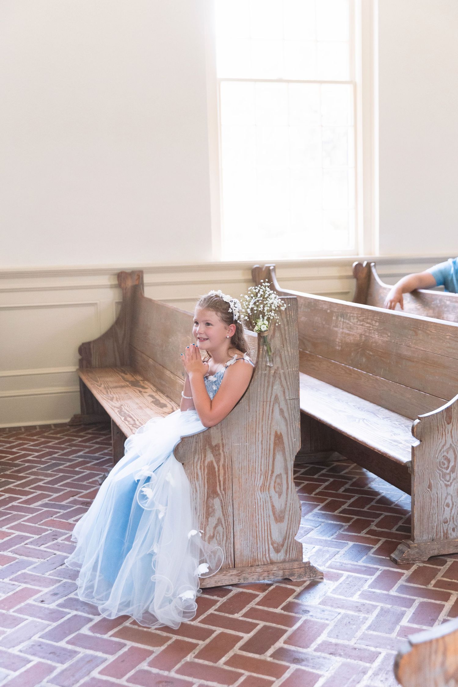 Adorable flower girl in blue dress sits on church pew with hands clasped at a wedding ceremony.