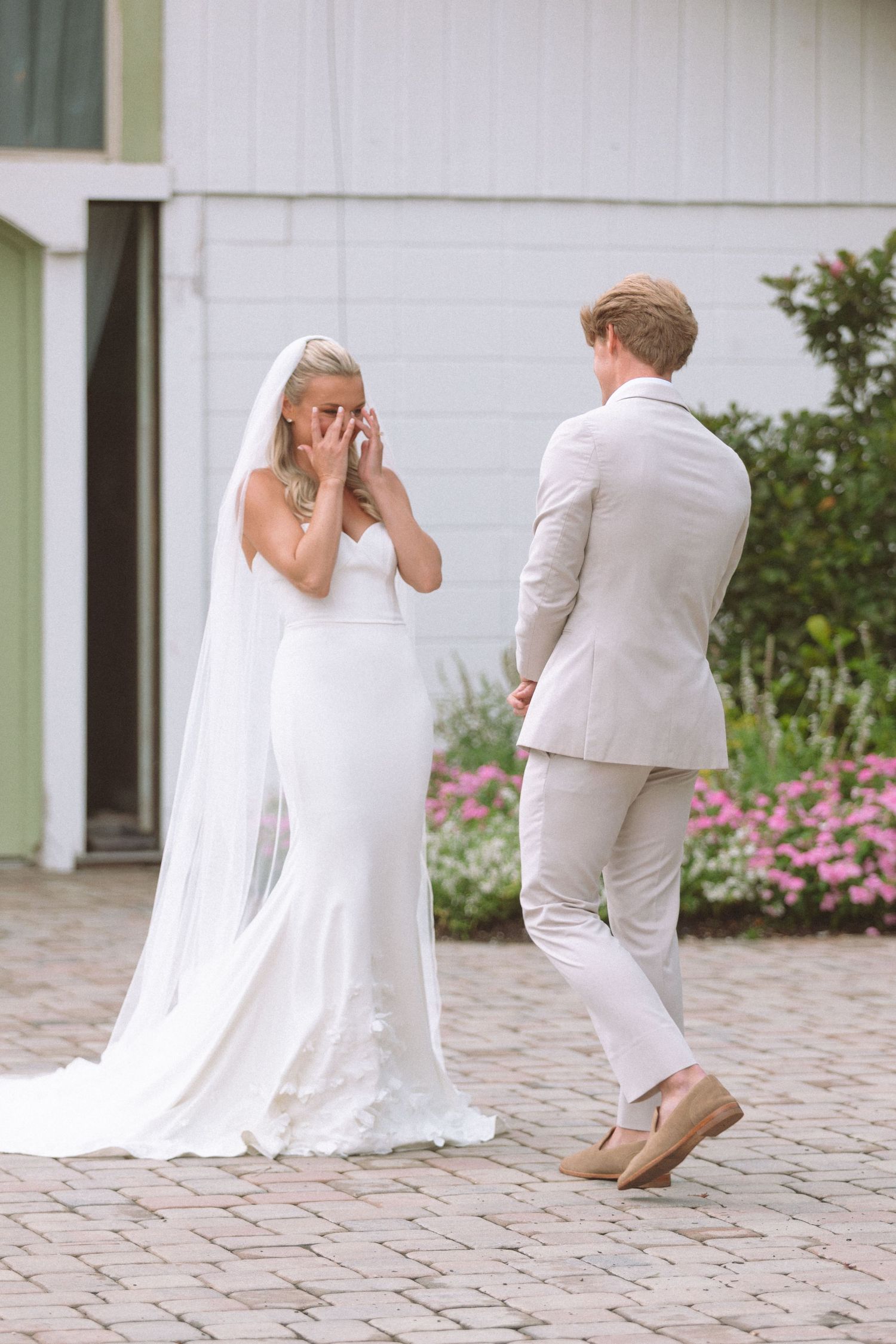 Emotional bride wiping tears during first look moment with groom outside white barn.
