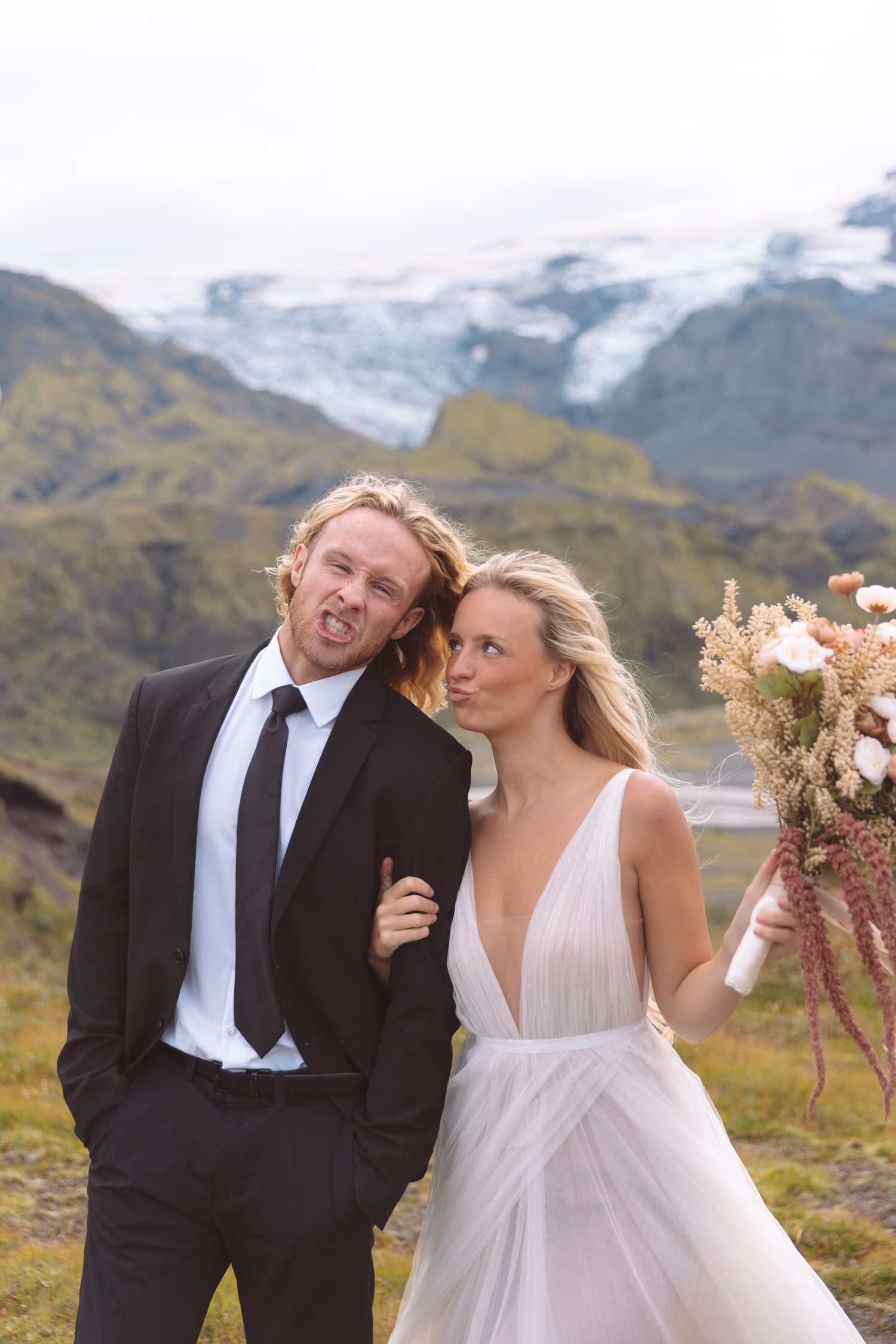 Smiling wedding couple pose playfully outdoors holding a bouquet with a glacier mountain landscape.
