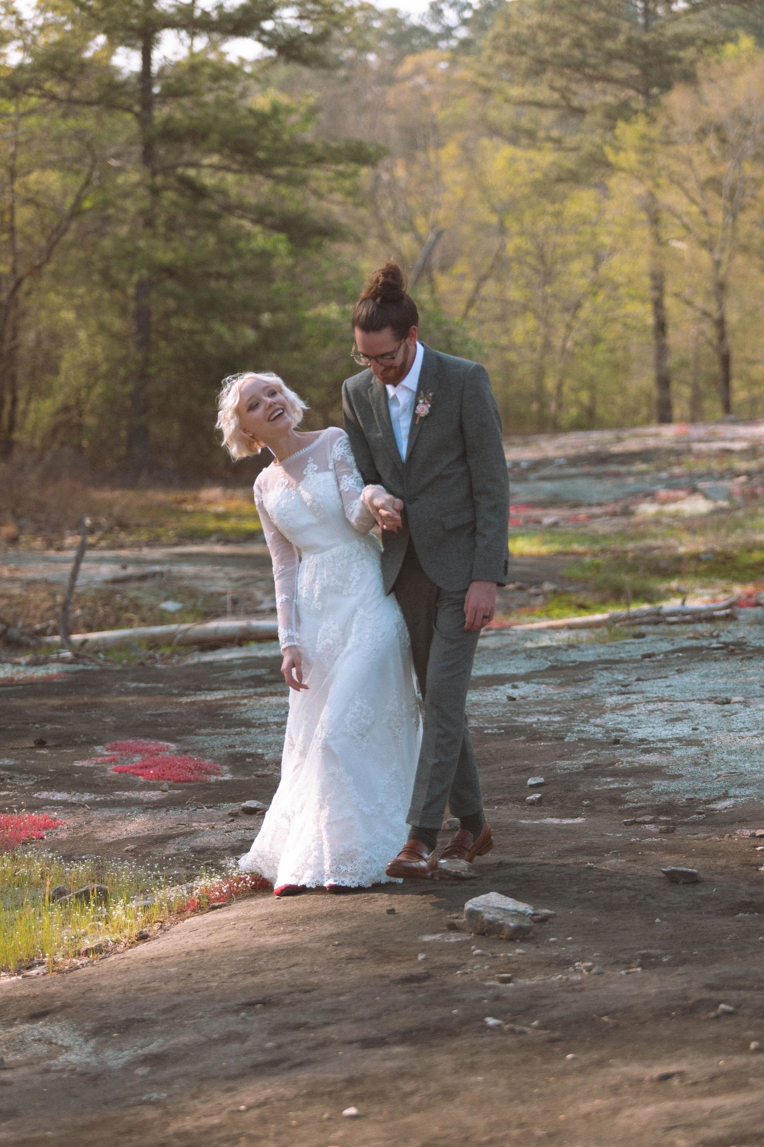 Bride in lace gown and groom in gray suit walking hand-in-hand on sun-drenched rocky landscape.