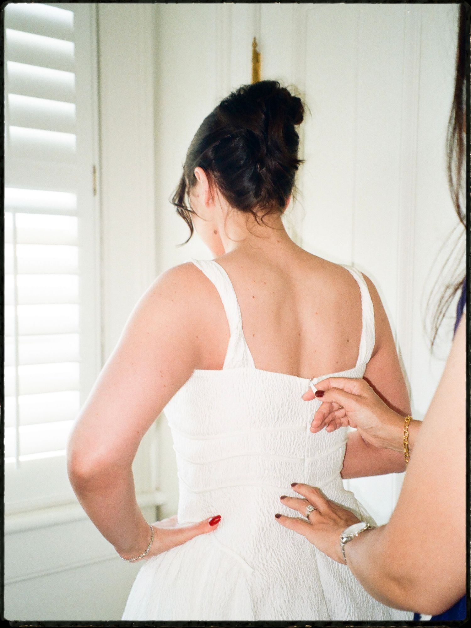 Bridesmaid fastening the back of a bride's white wedding dress near white shuttered windows.