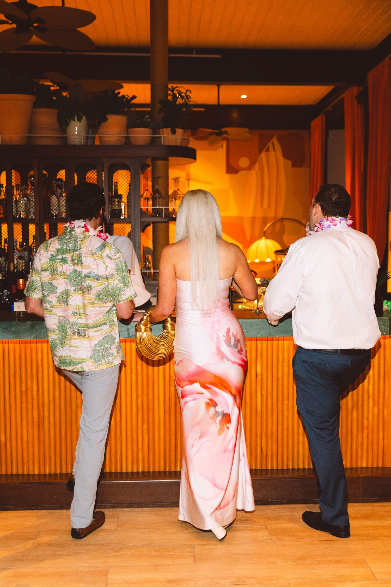 Three guests in tropical attire standing at a warmly lit tiki-style bar during a wedding reception.