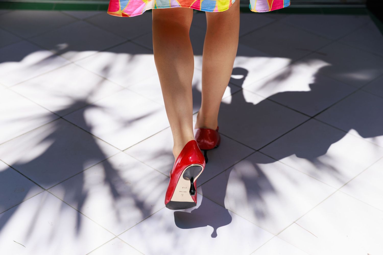 Woman wearing red high heels and colorful dress walking on tiled floor with palm tree shadows.