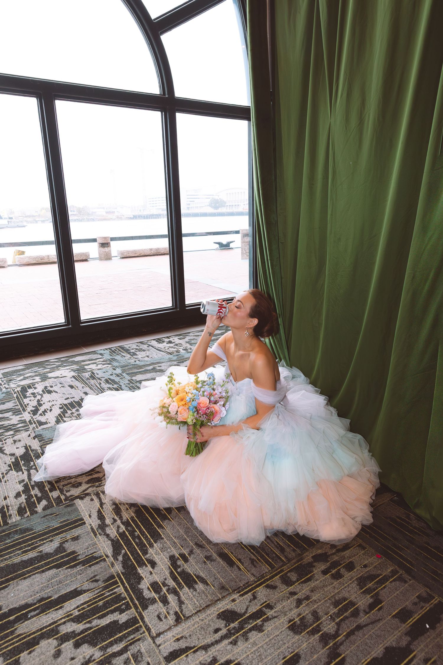 Bride in colorful pastel tulle gown sits on floor holding bouquet near large window with green curtains.