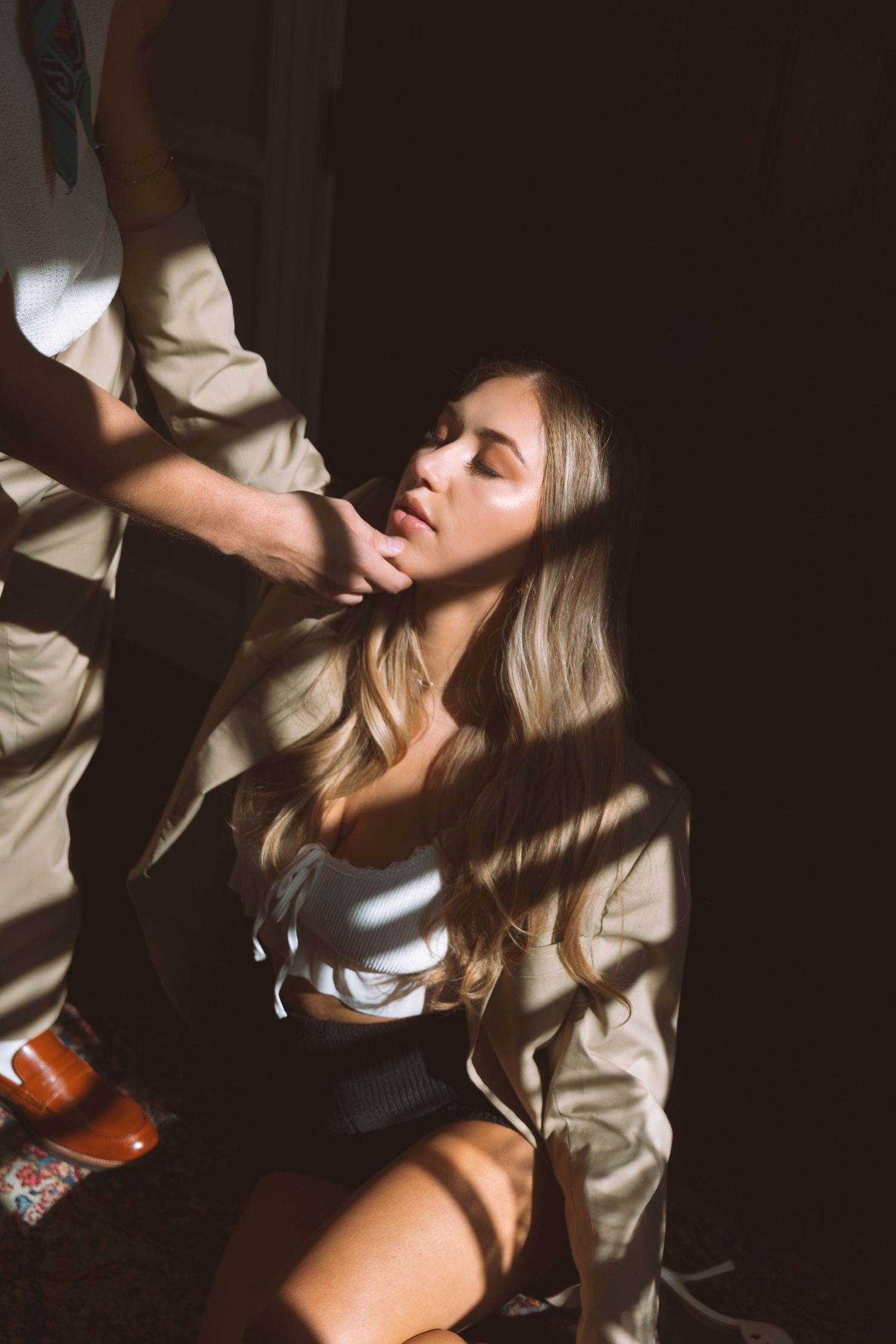 Blonde woman in beige jacket sitting on floor with sunlight streaming through dark shadows.