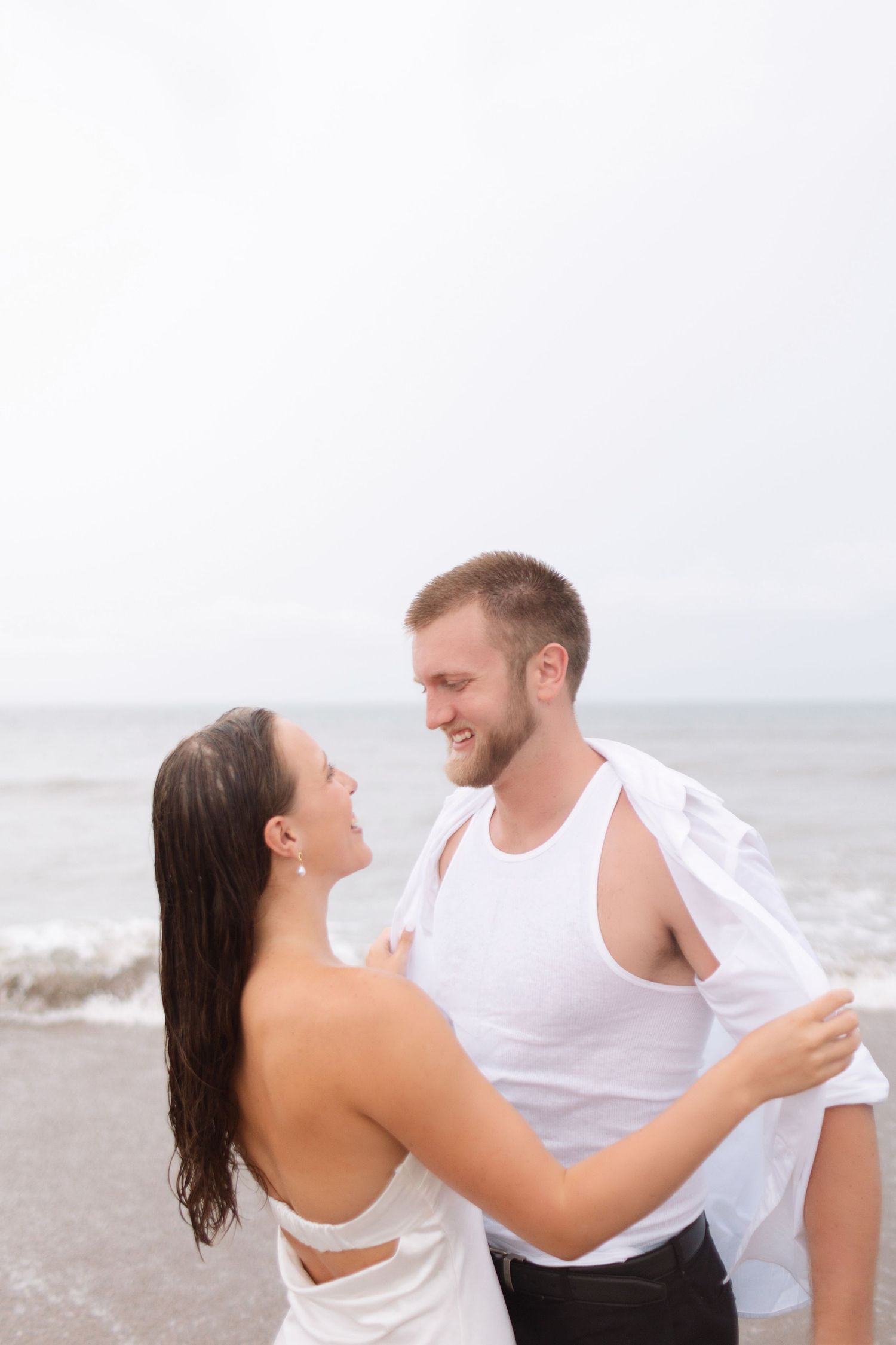Happy couple in white outfits embracing and smiling at each other on a cloudy beach.