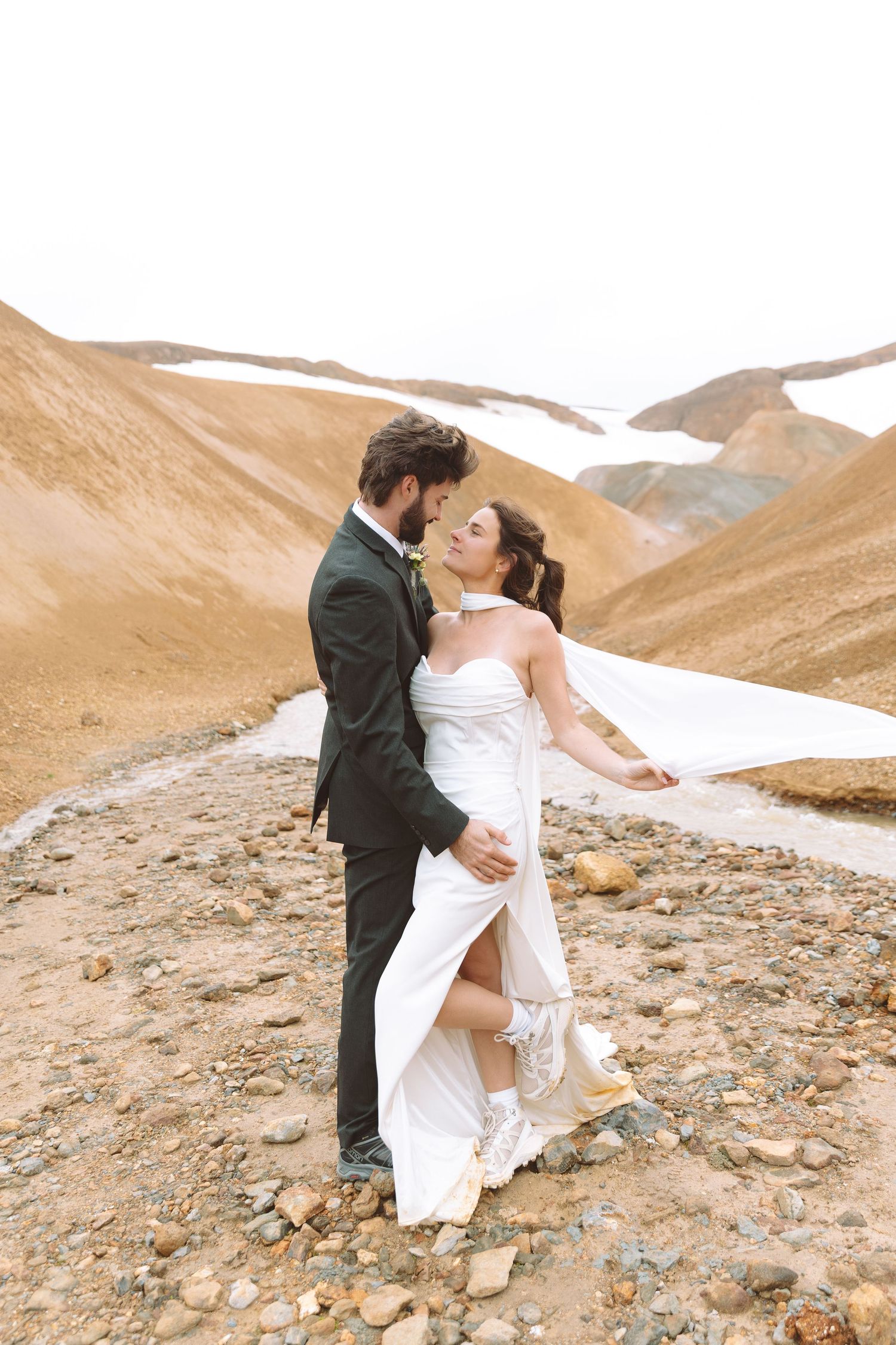 Wedding couple embraces on rocky terrain with dramatic brown mountains and snow in the background.