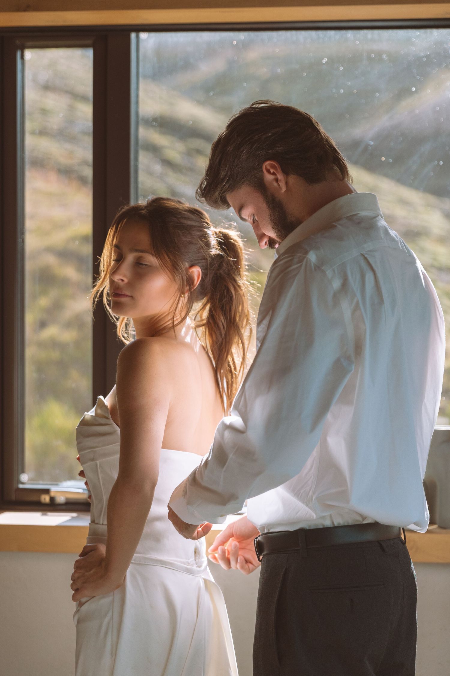 Groom tenderly holding brides hands from behind in warm golden window light.