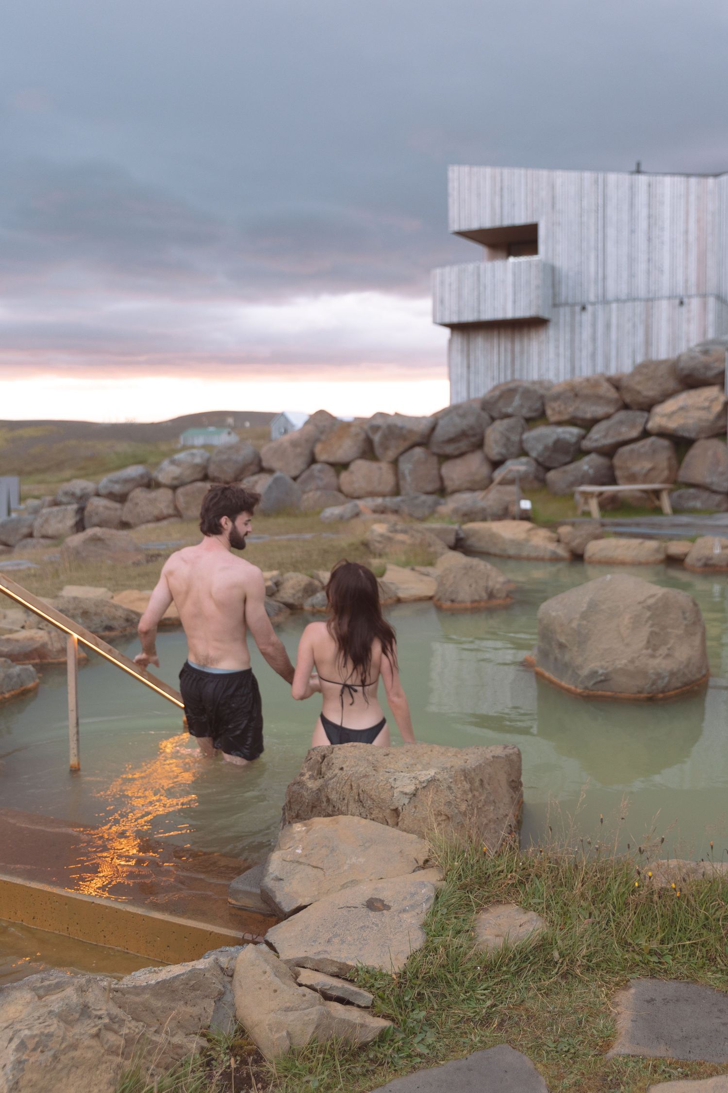 Couple holding hands entering a natural geothermal hot spring pool at dusk with dramatic cloudy sky in Iceland.