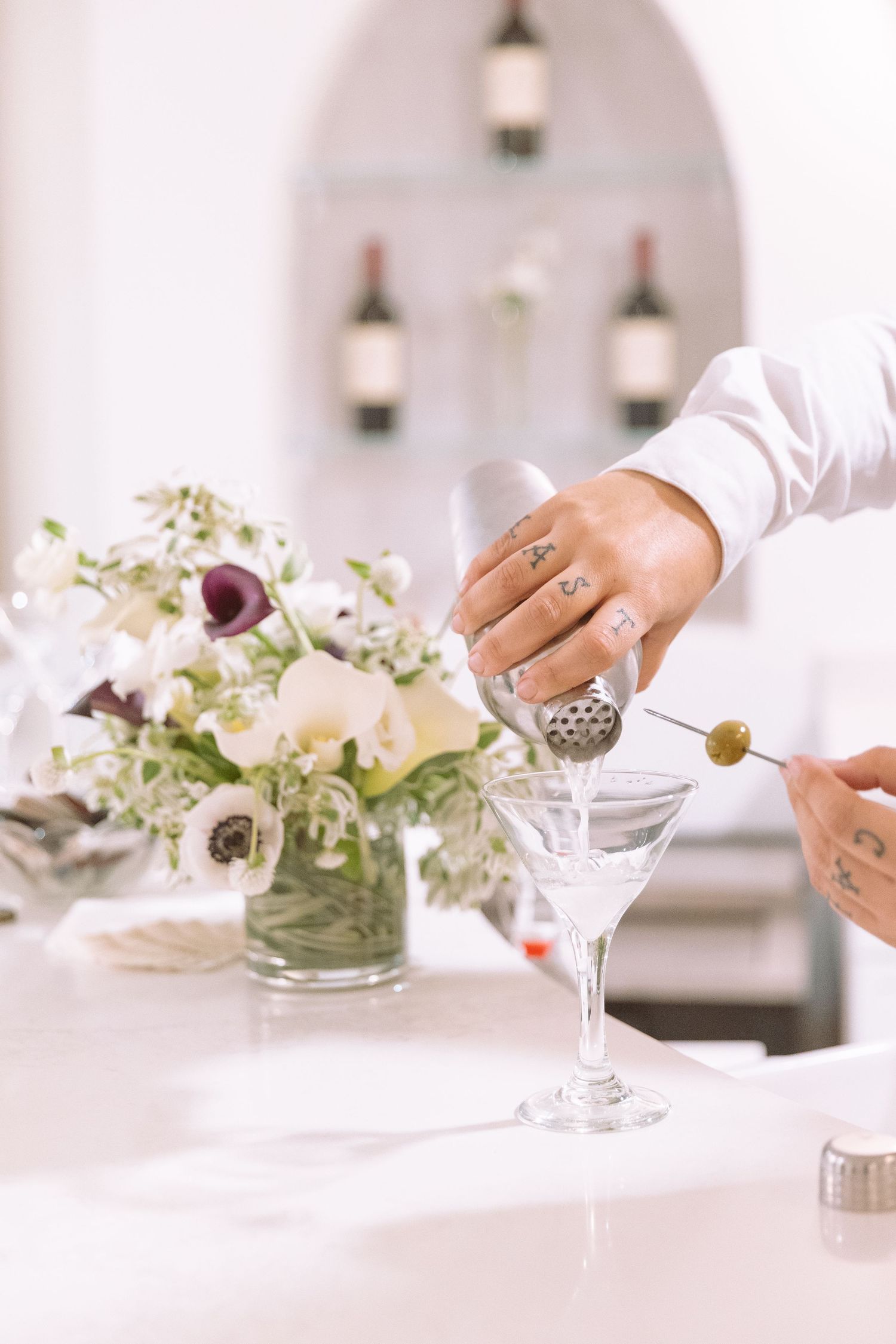 Tattooed bartender preparing a dirty martini with olive beside a white floral arrangement.