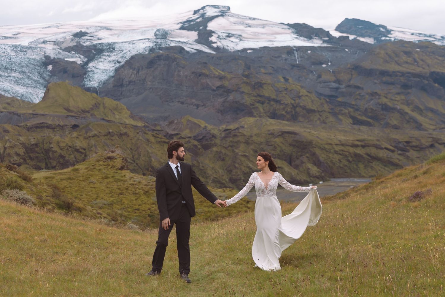 Couple holding hands in wedding attire with glacial mountains in Iceland backdrop.
