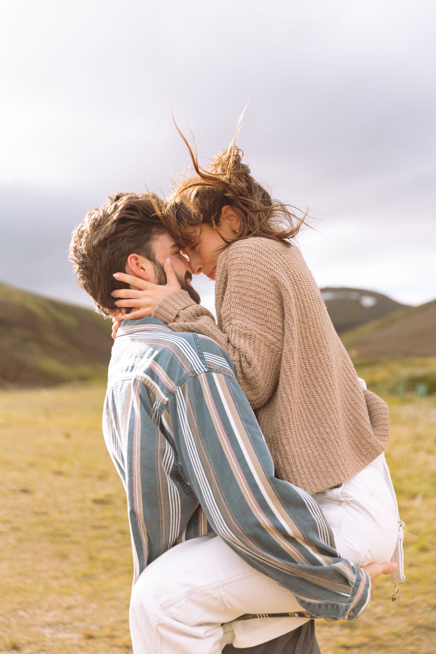 Couple sharing a romantic kiss while man lifts woman in a scenic green mountain meadow.