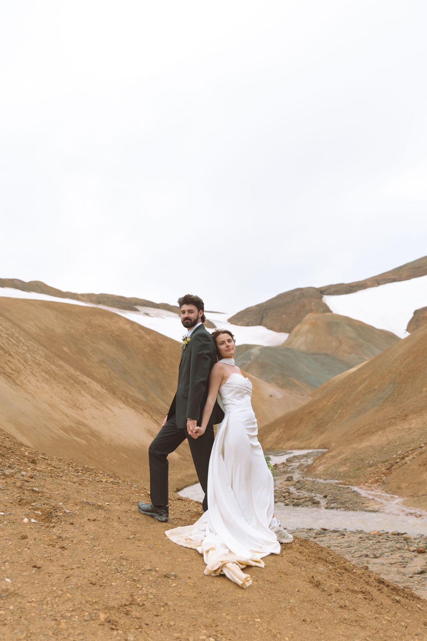 Bride and groom hold hands posing back-to-back on a rugged mountain trail during their outdoor wedding.
