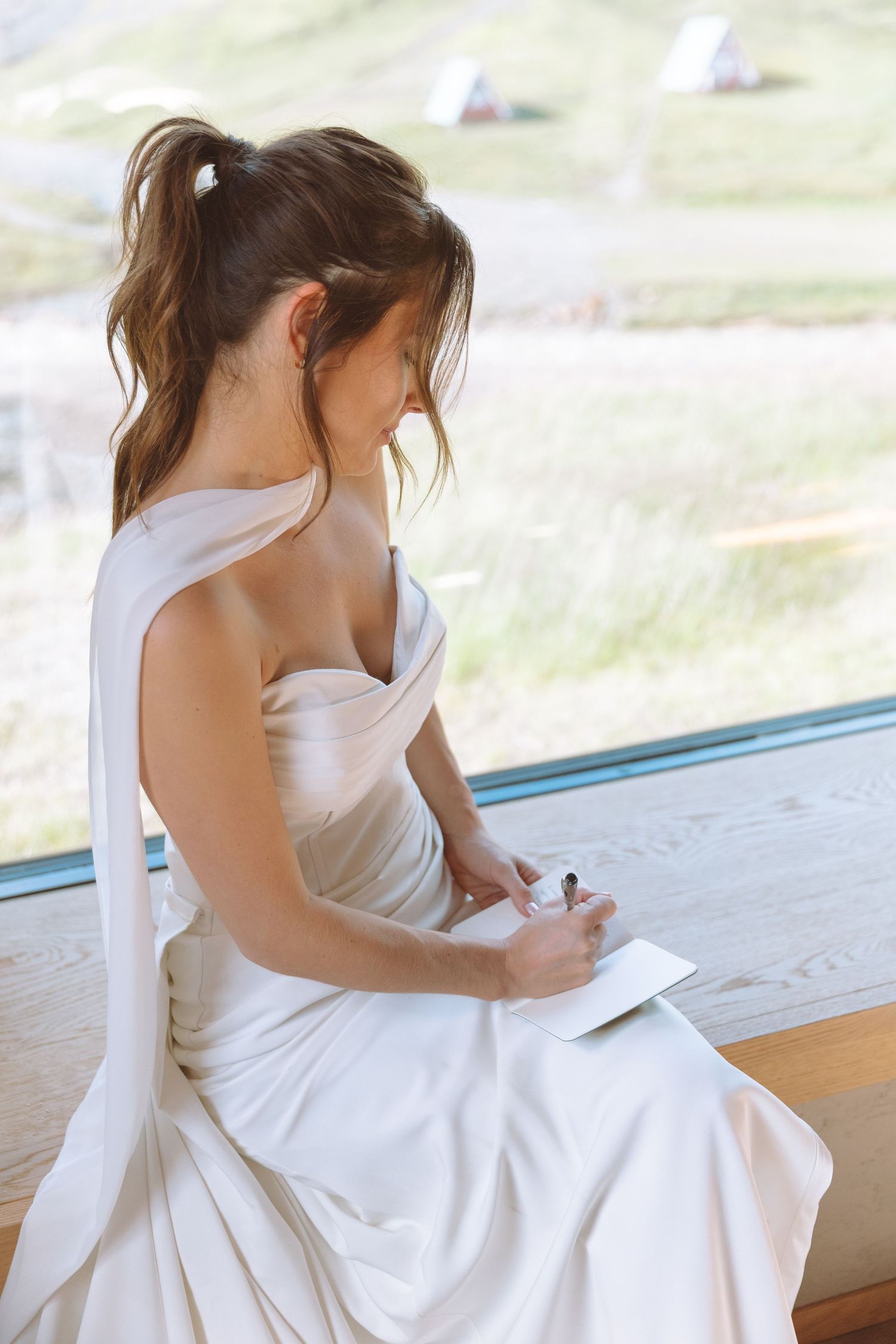 Elegant bride in strapless white gown writing wedding vows in notebook by large window.