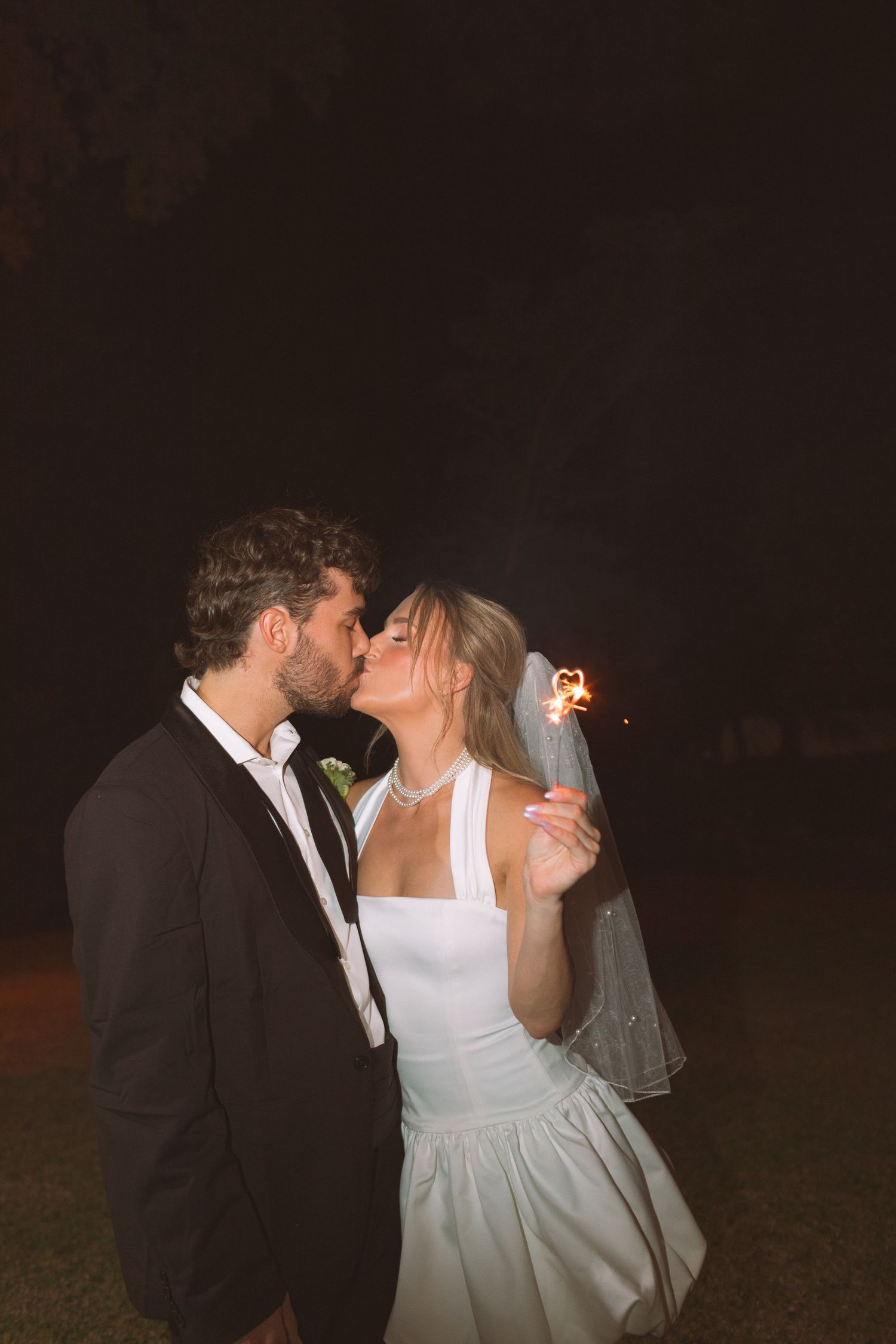 Bride in white mini dress and veil kissing groom while holding sparkler during nighttime wedding celebration.